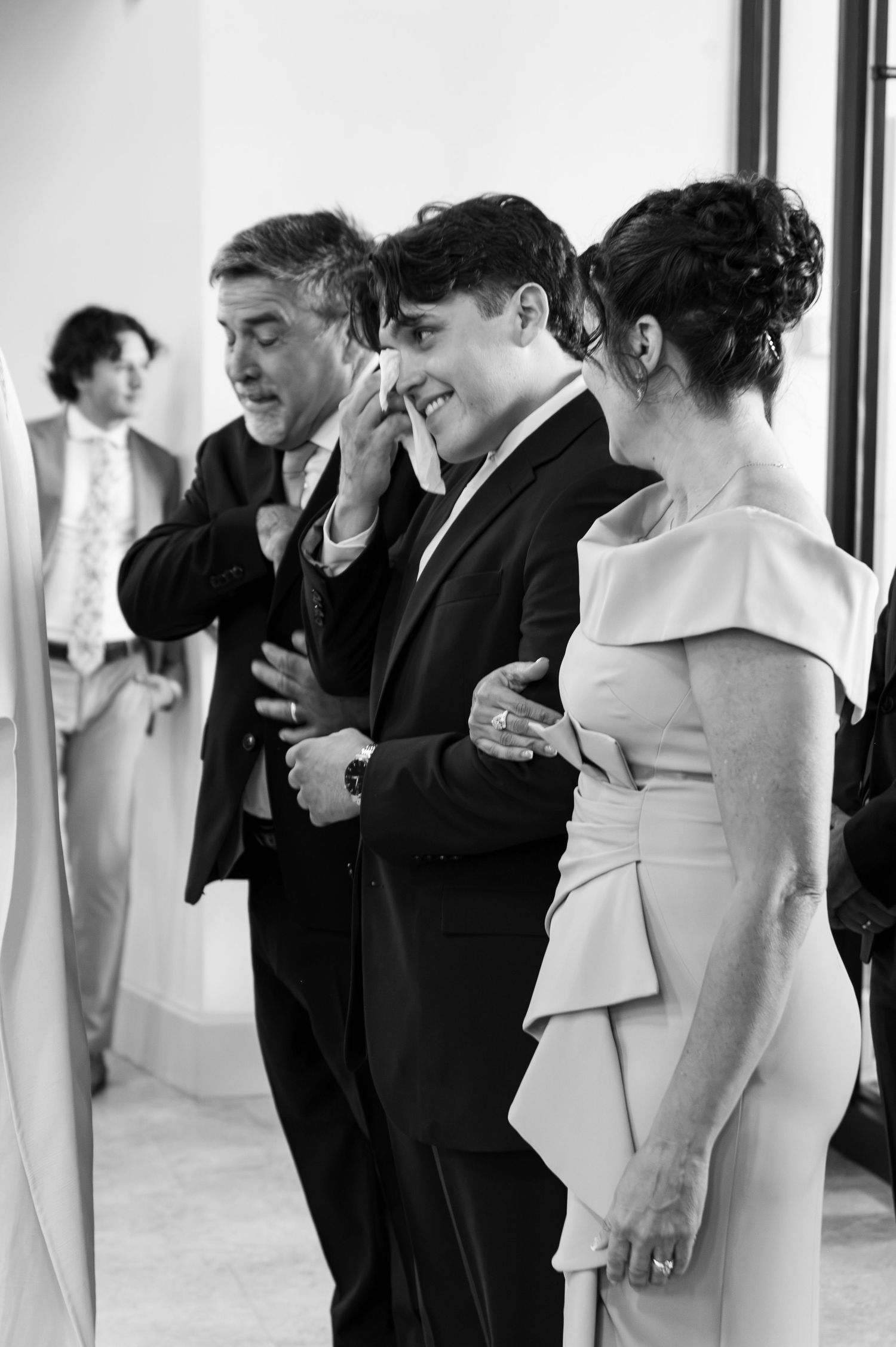 Emotional groom wiping tears with parents during wedding day first look moment.