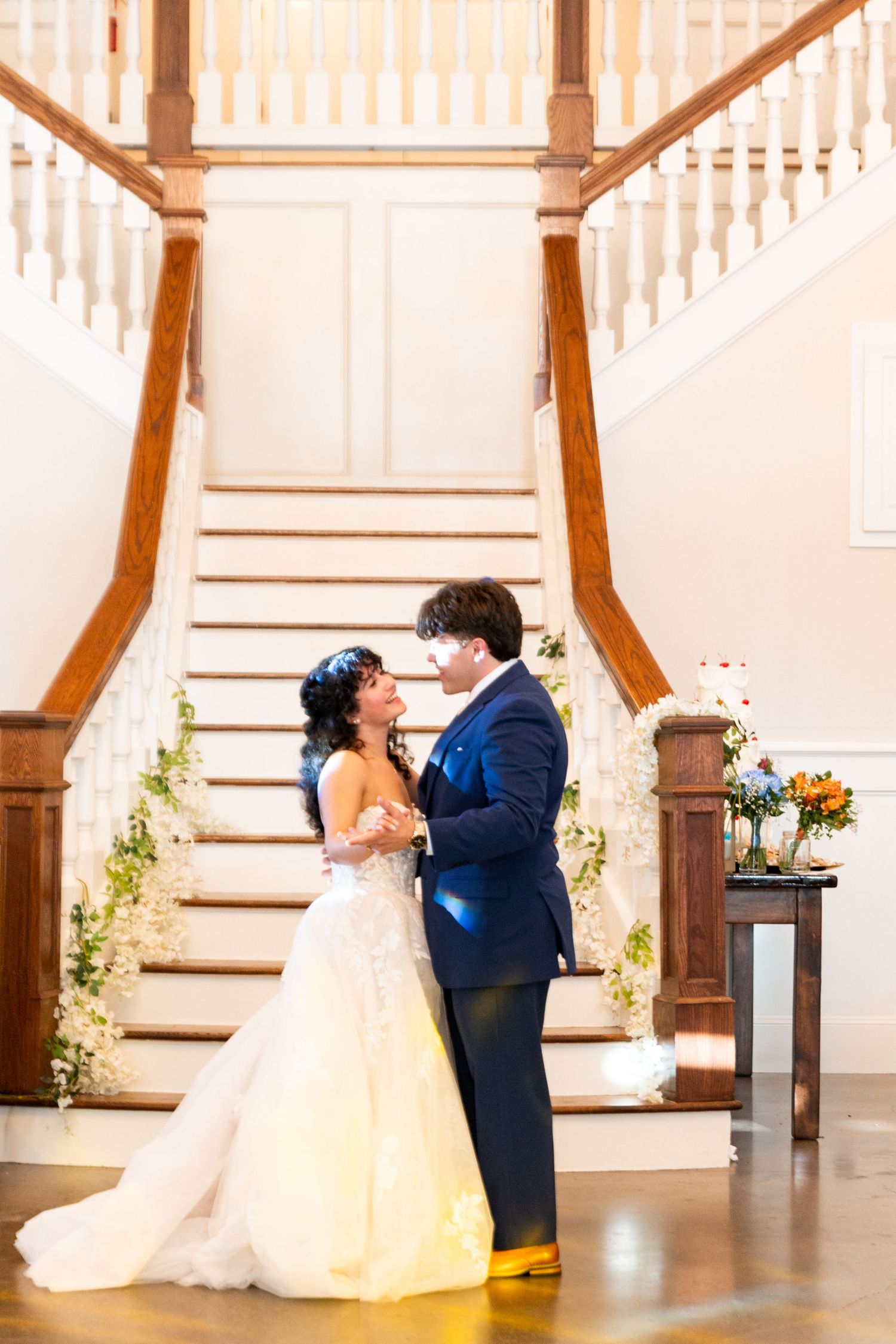 Bride and groom share first dance before elegant staircase decorated with florals at wedding reception.
