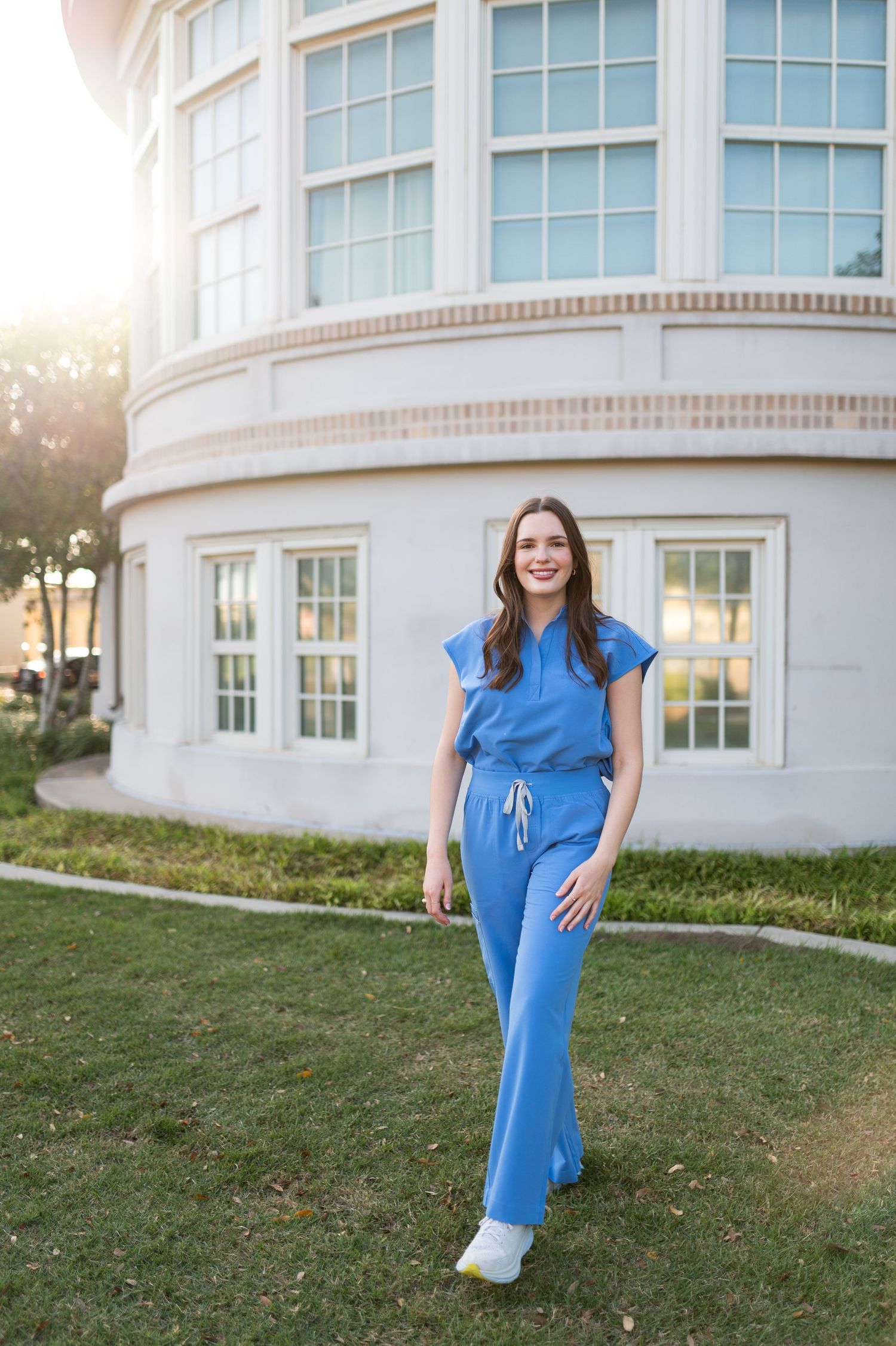 Young woman in blue matching set outfit poses smiling on green grass in warm evening sunlight.