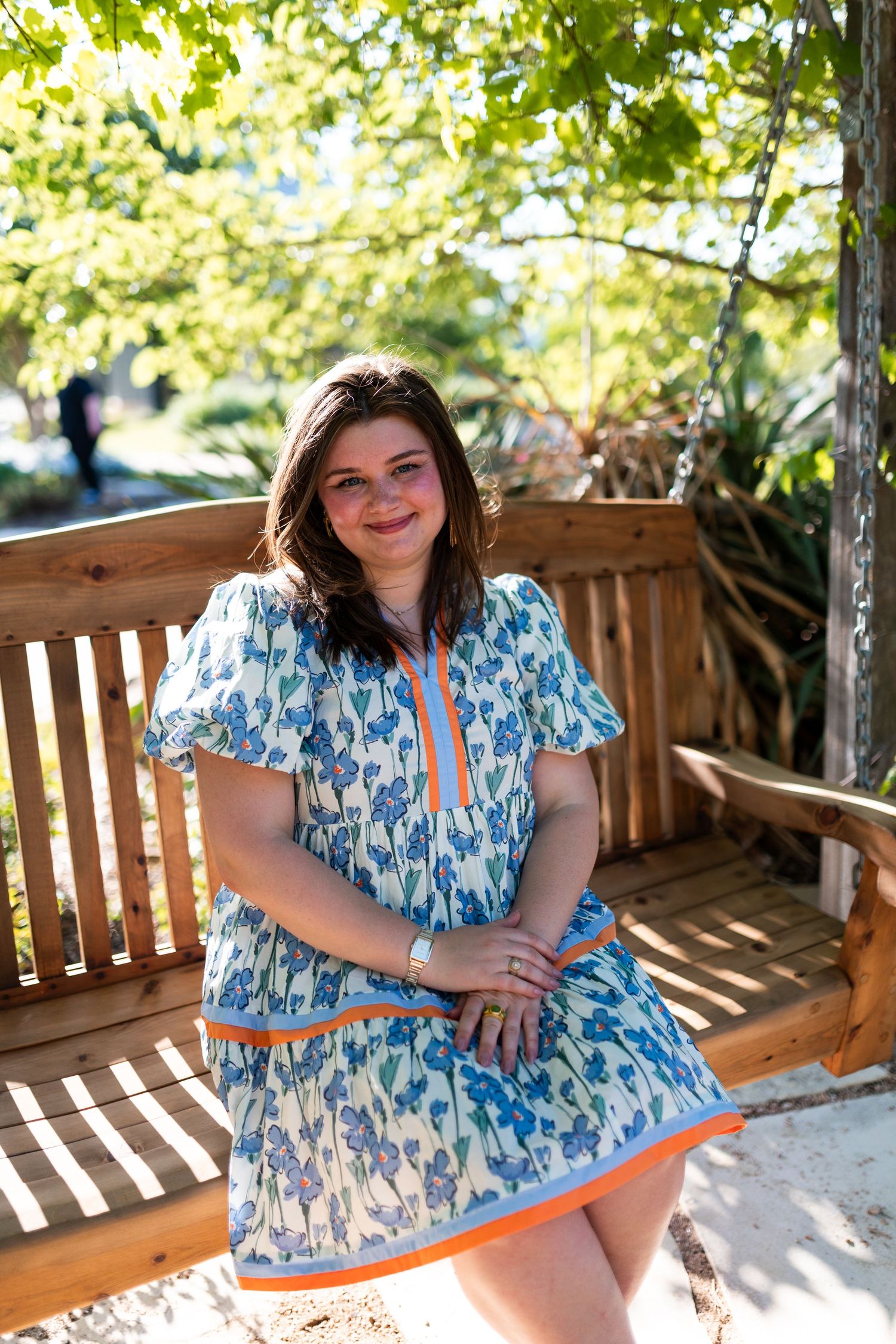 Smiling woman in blue floral dress with orange trim sitting on wooden bench outdoors.