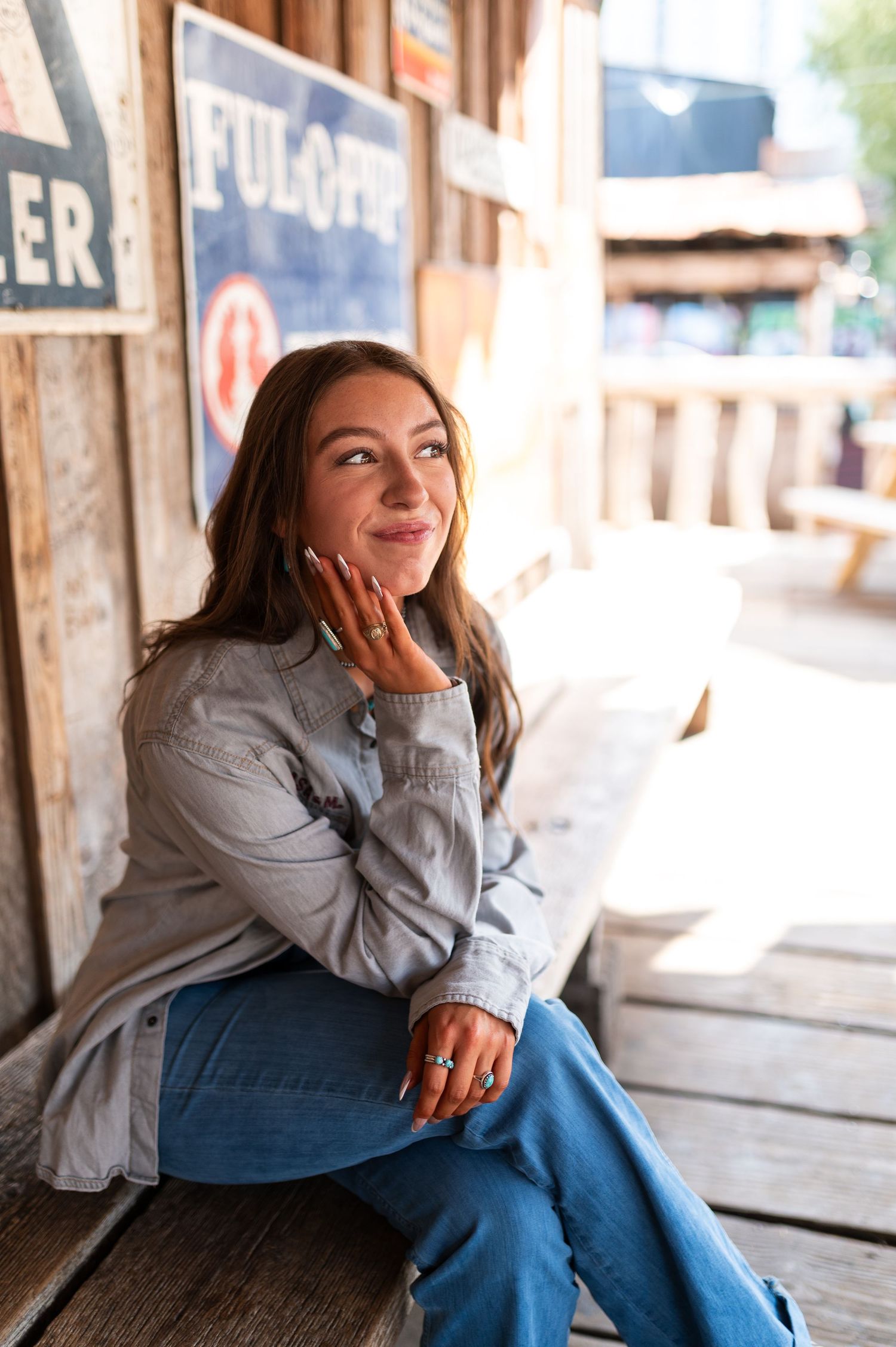 Smiling young woman in denim jacket sits on rustic wooden porch with vintage signs behind her.