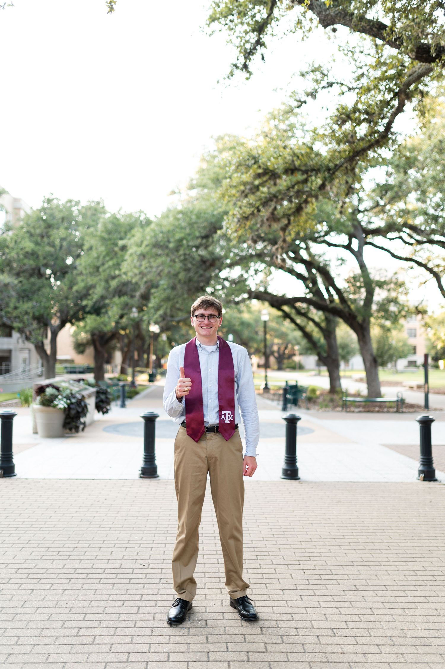 Smiling Texas A&M graduate in maroon tie and stole gives thumbs up on campus brick walkway.