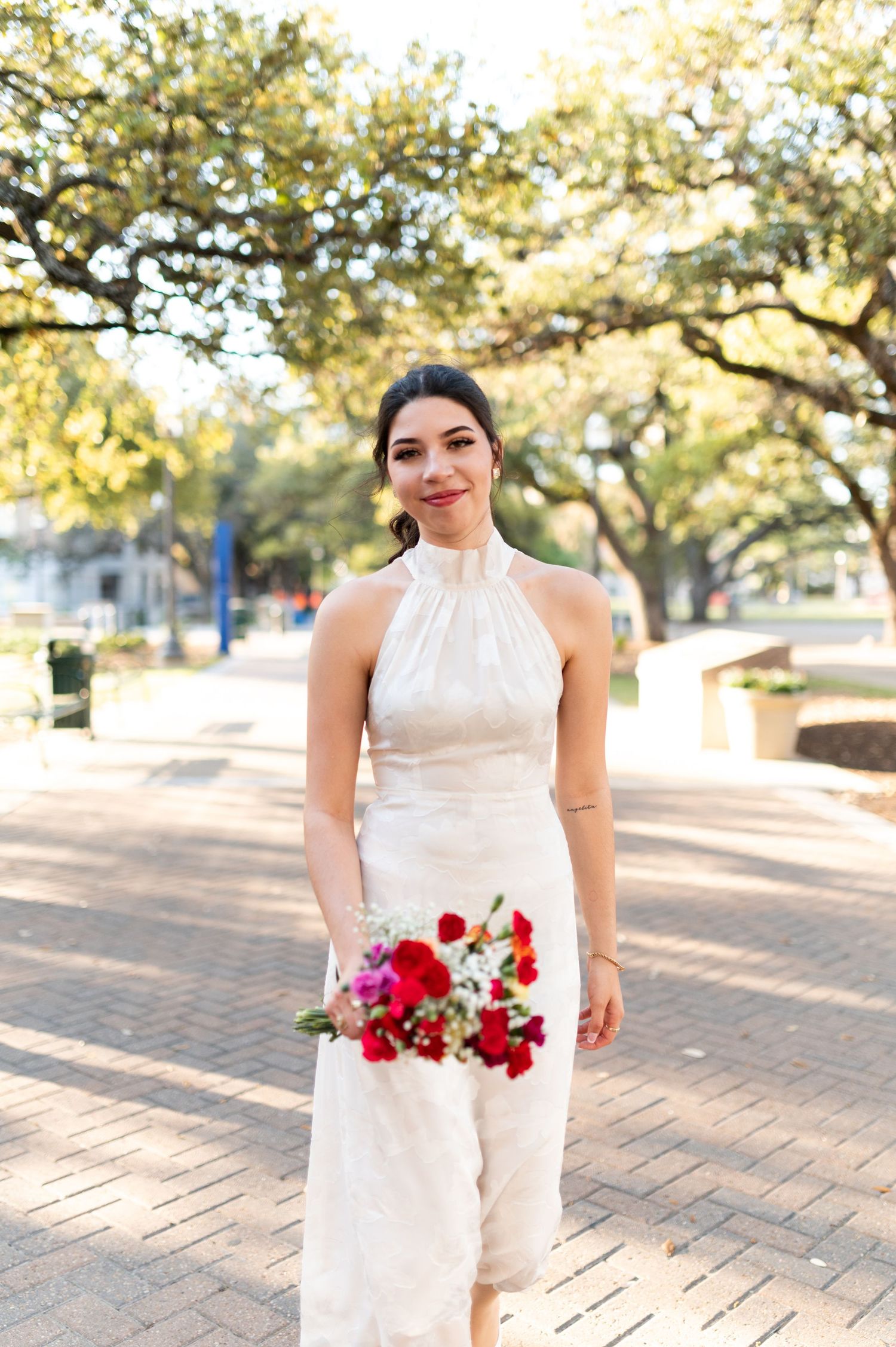 Elegant bride in white halter gown holds colorful red bouquet while standing on brick pathway under oak trees.