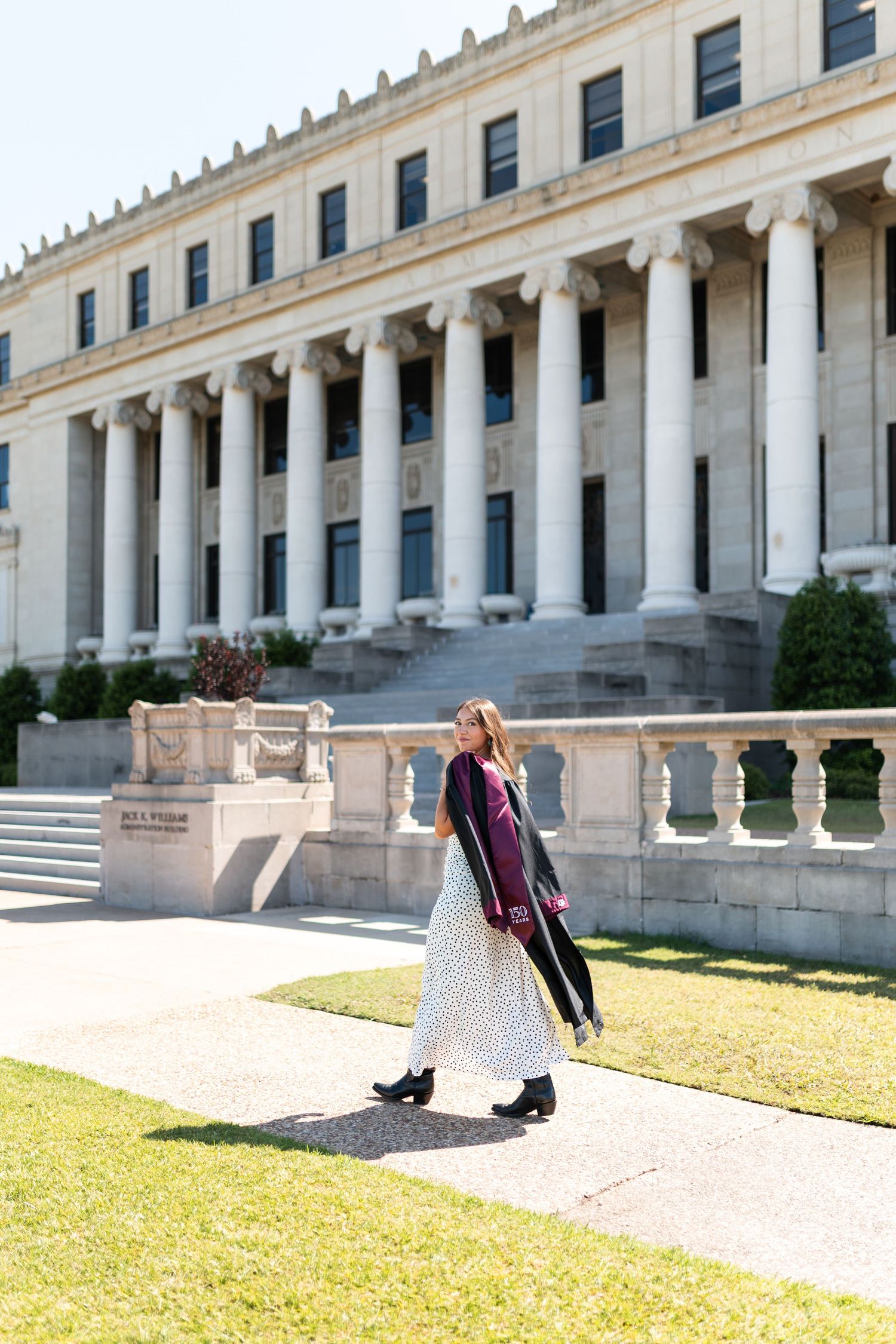 Texas A&M graduate walks across campus lawn in black graduation robe and maroon stole before neoclassical building. Grad Photos at the Admin