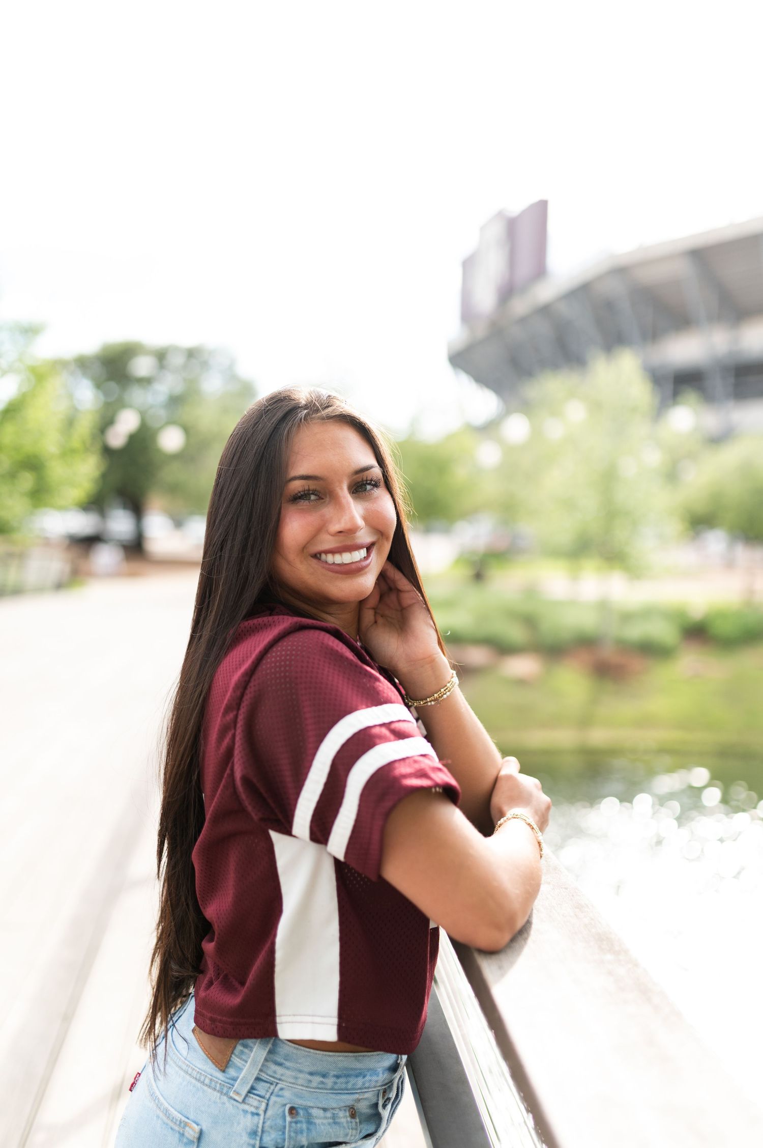 Smiling young woman in maroon Texas A&M jersey leaning on railing with stadium in background.
