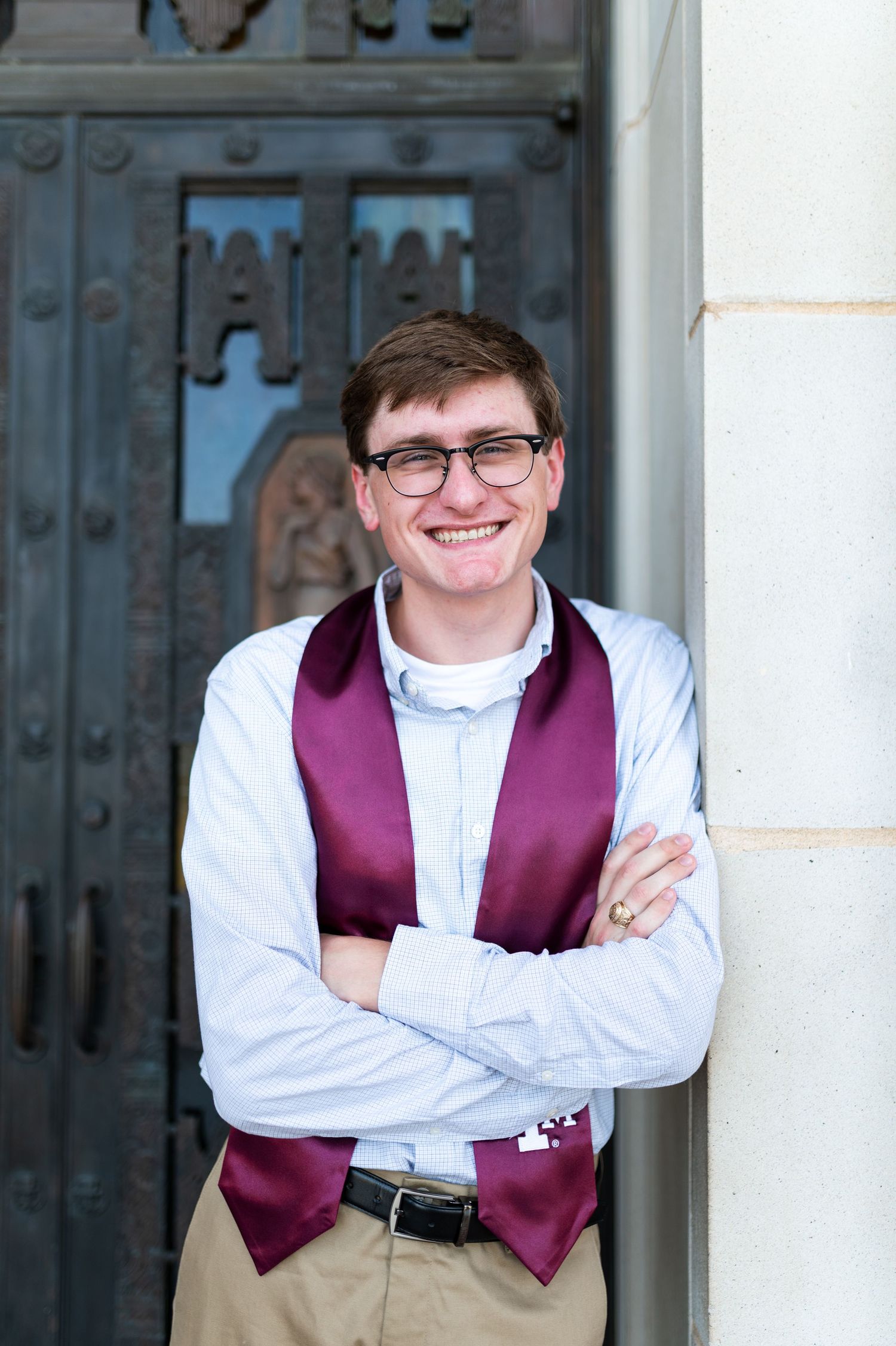 Smiling male Texas A&M graduate with glasses and maroon stole leans against ornate campus gate. Grad Photos at the Texas A&M Admin building