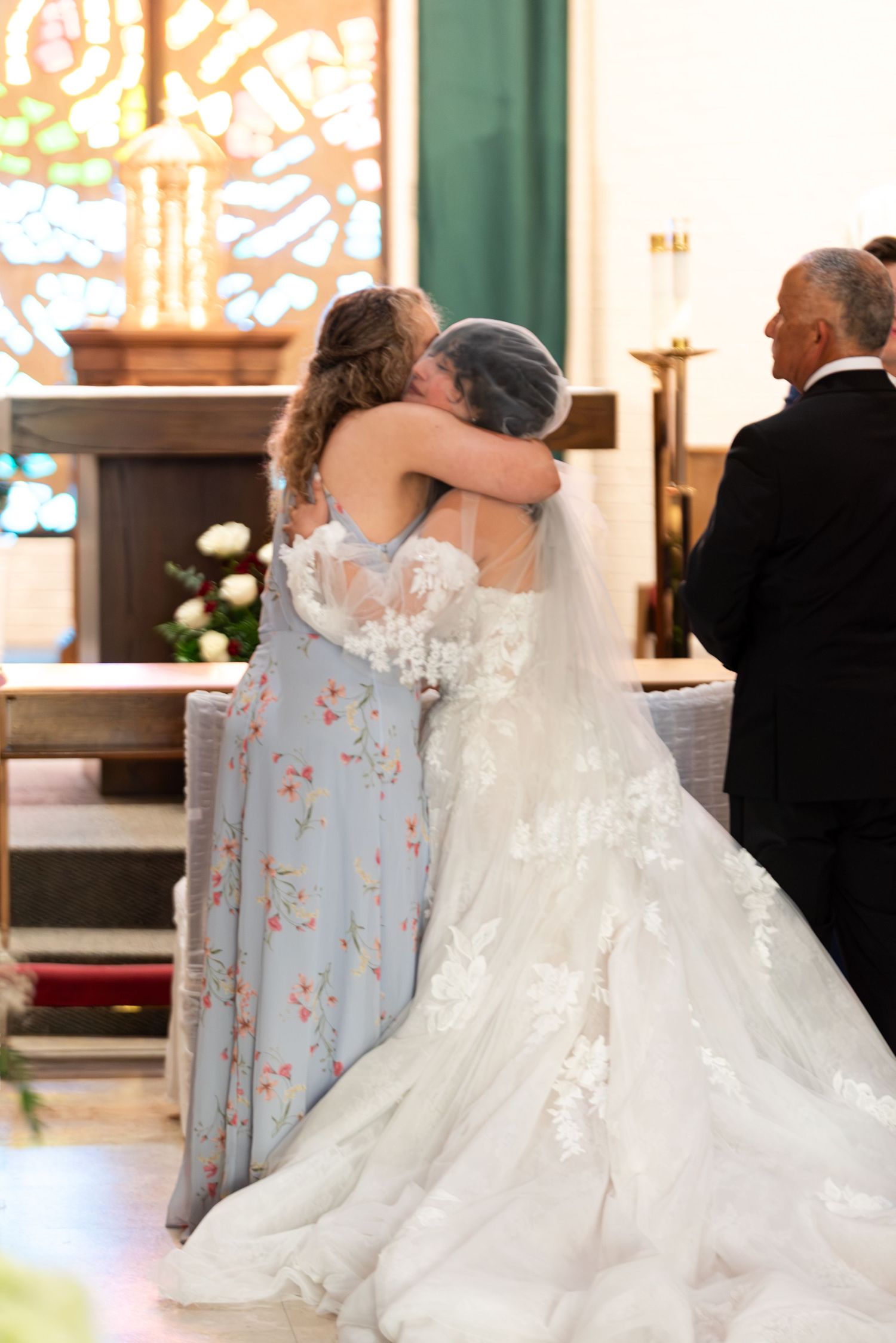 Bride in lace gown embraces woman in floral blue dress during emotional church wedding ceremony moment.