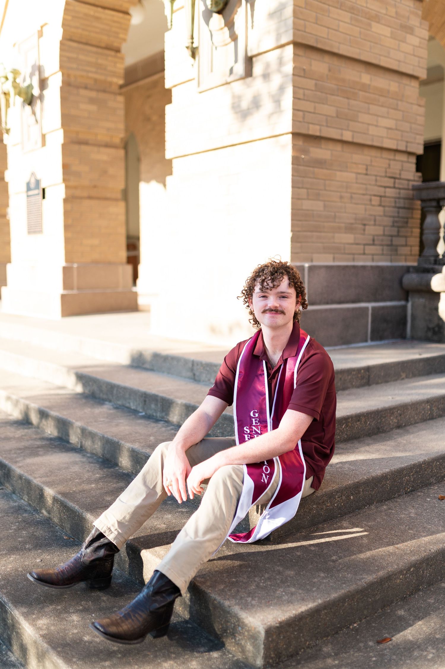 Young man in maroon shirt with graduation stole sits smiling on stone steps.