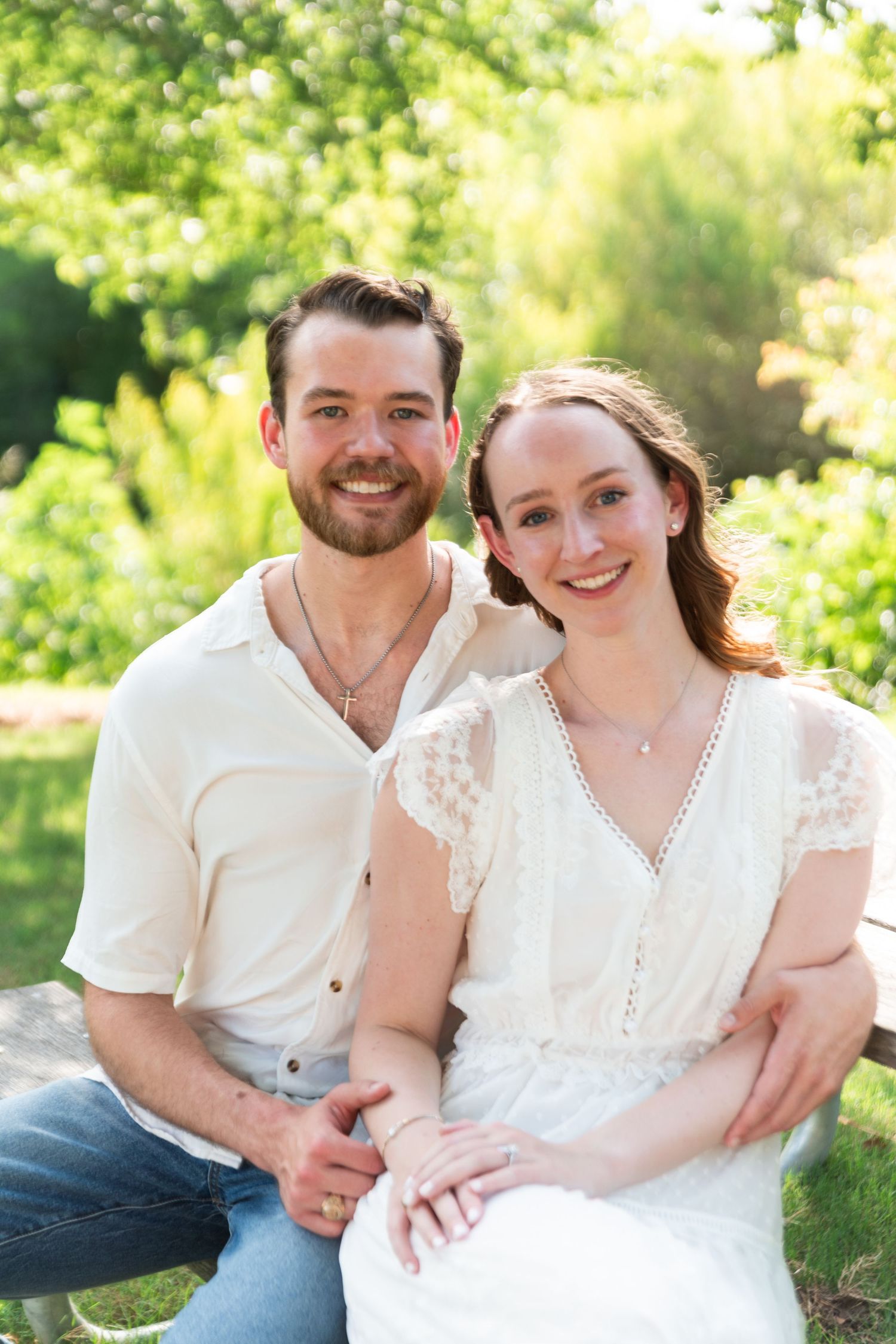 A smiling couple sits together on a bench in a sunny green garden, dressed in casual white and cream outfits.