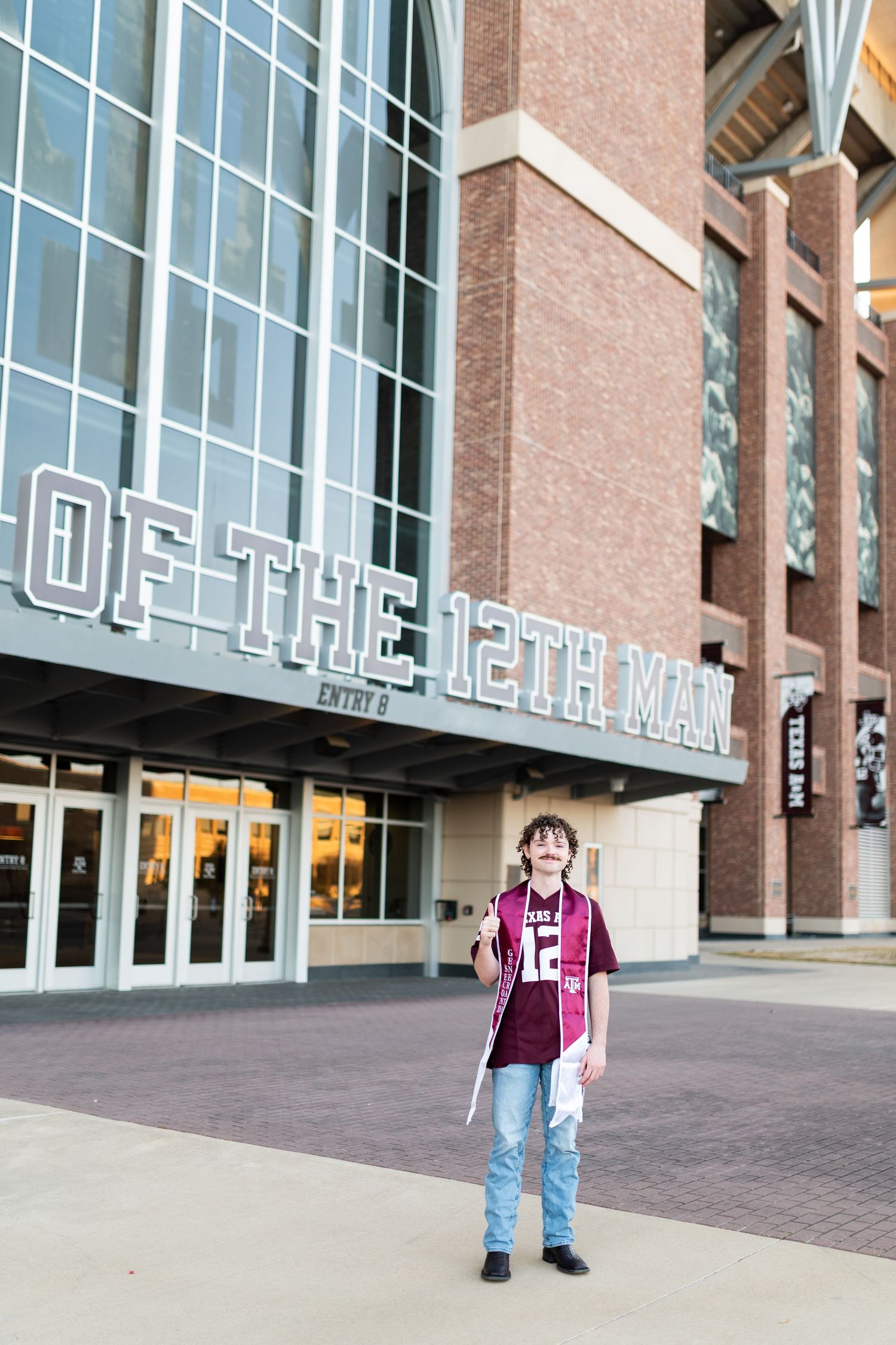 Texas A&M graduate in maroon jersey and stole stands outside Of The 12th Man stadium entrance.