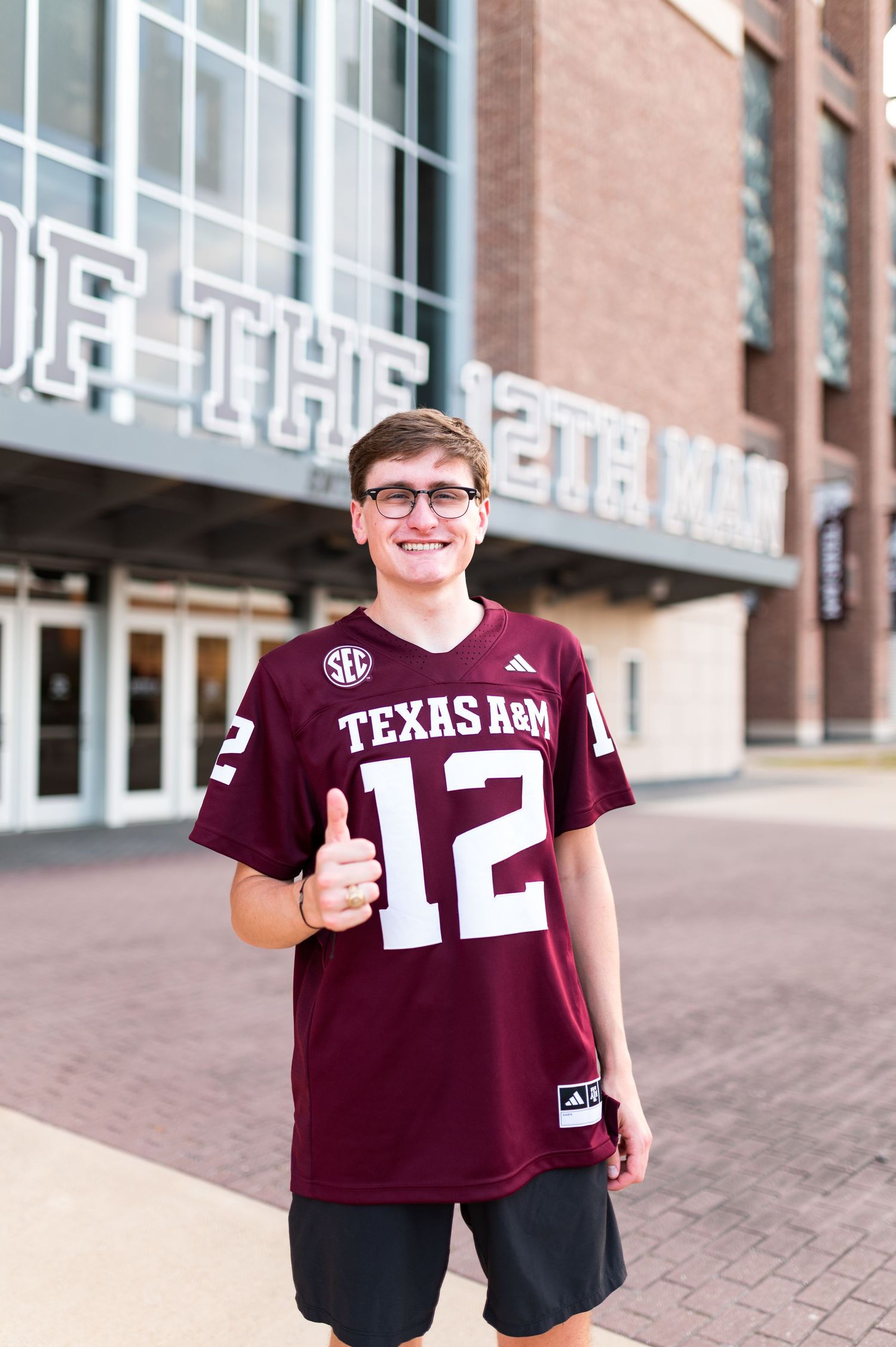 Young man in Texas A&M number 12 jersey gives thumbs up outside Kyle Field stadium.