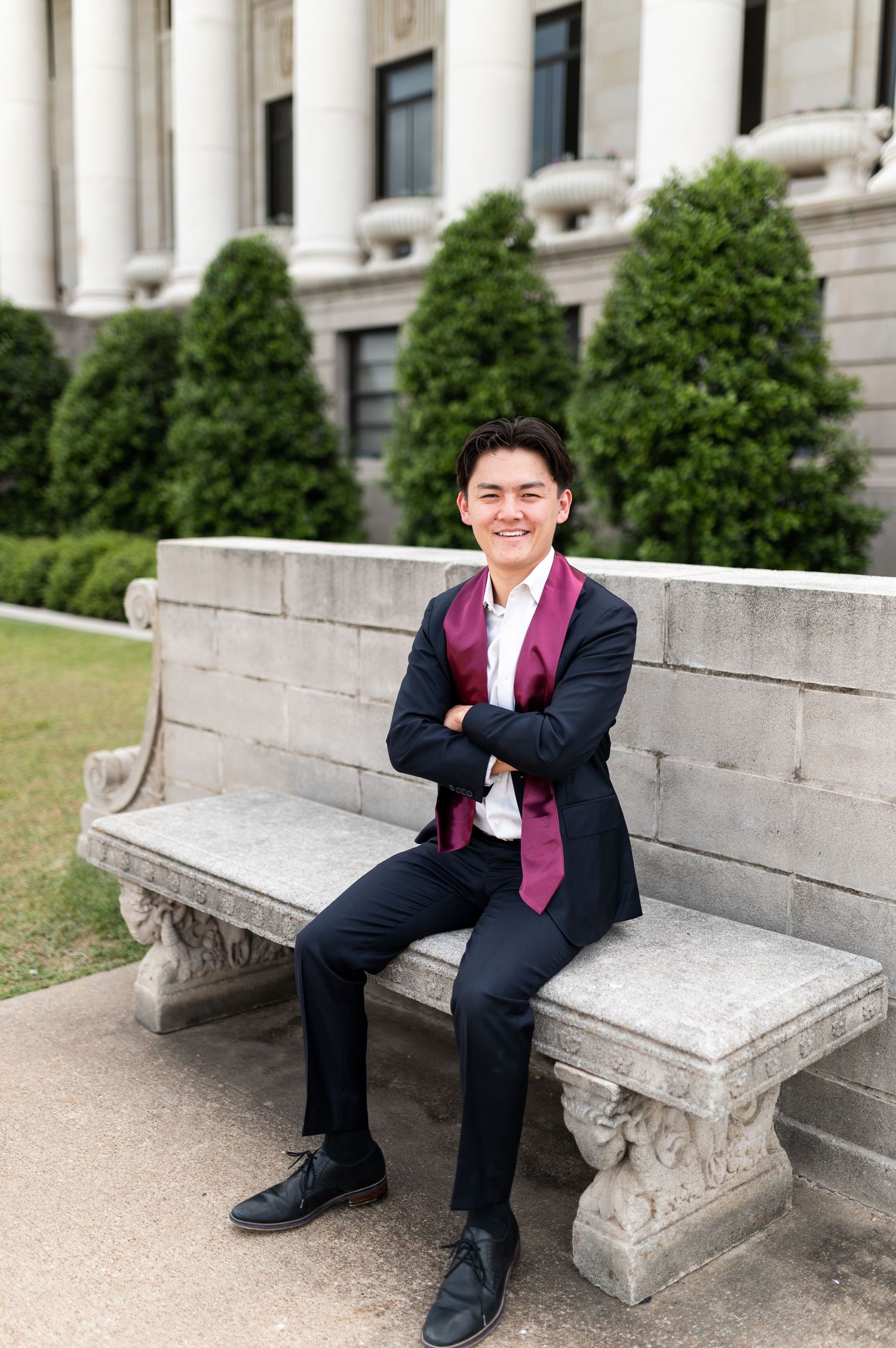 Young man in navy suit with maroon graduation stole sitting cross-armed on stone bench outside university building. Grad Photos at the Admin