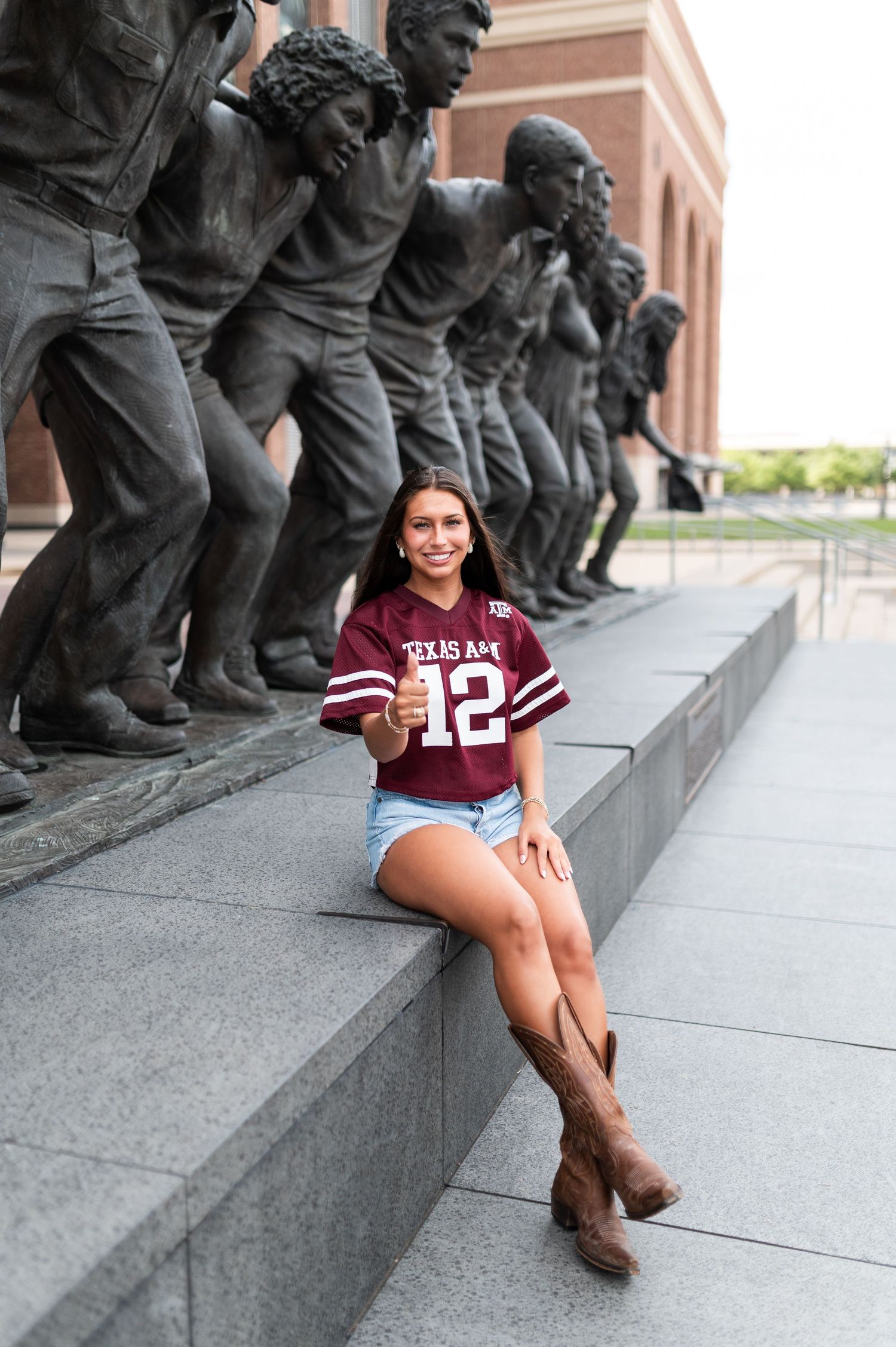 Smiling woman in Texas A&M jersey sits beside bronze statues outside Kyle Field.