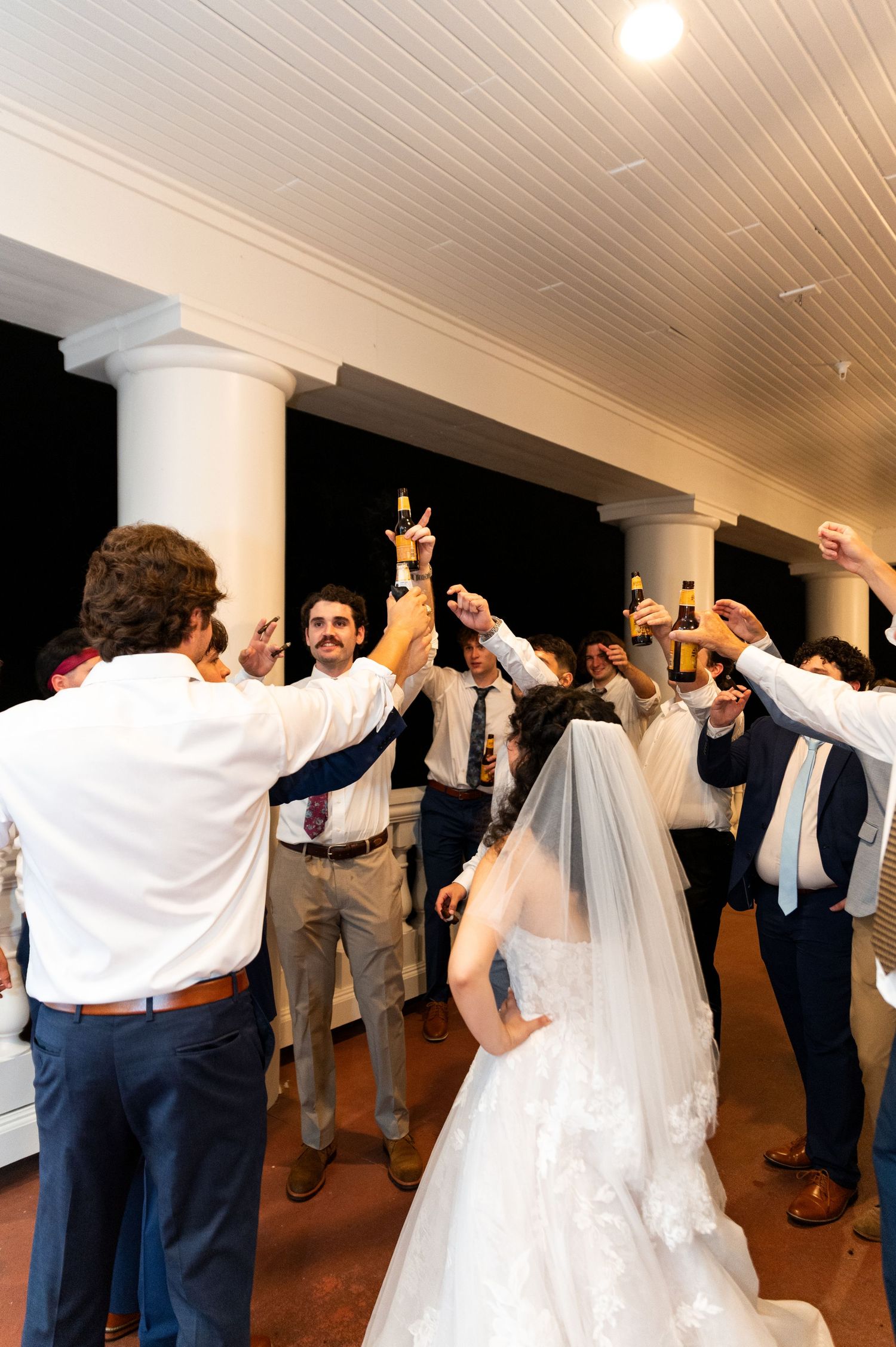 Wedding guests raising bottles in toast surrounding bride in lace gown on venue porch at night.