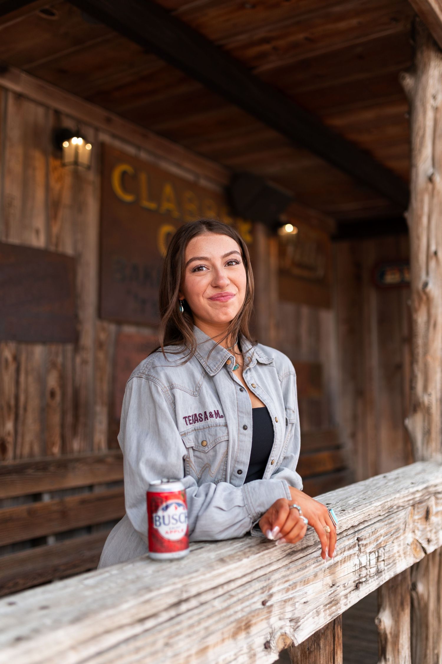 Smiling woman leaning on wooden rail with Busch beer can at rustic Clabber Girl building.