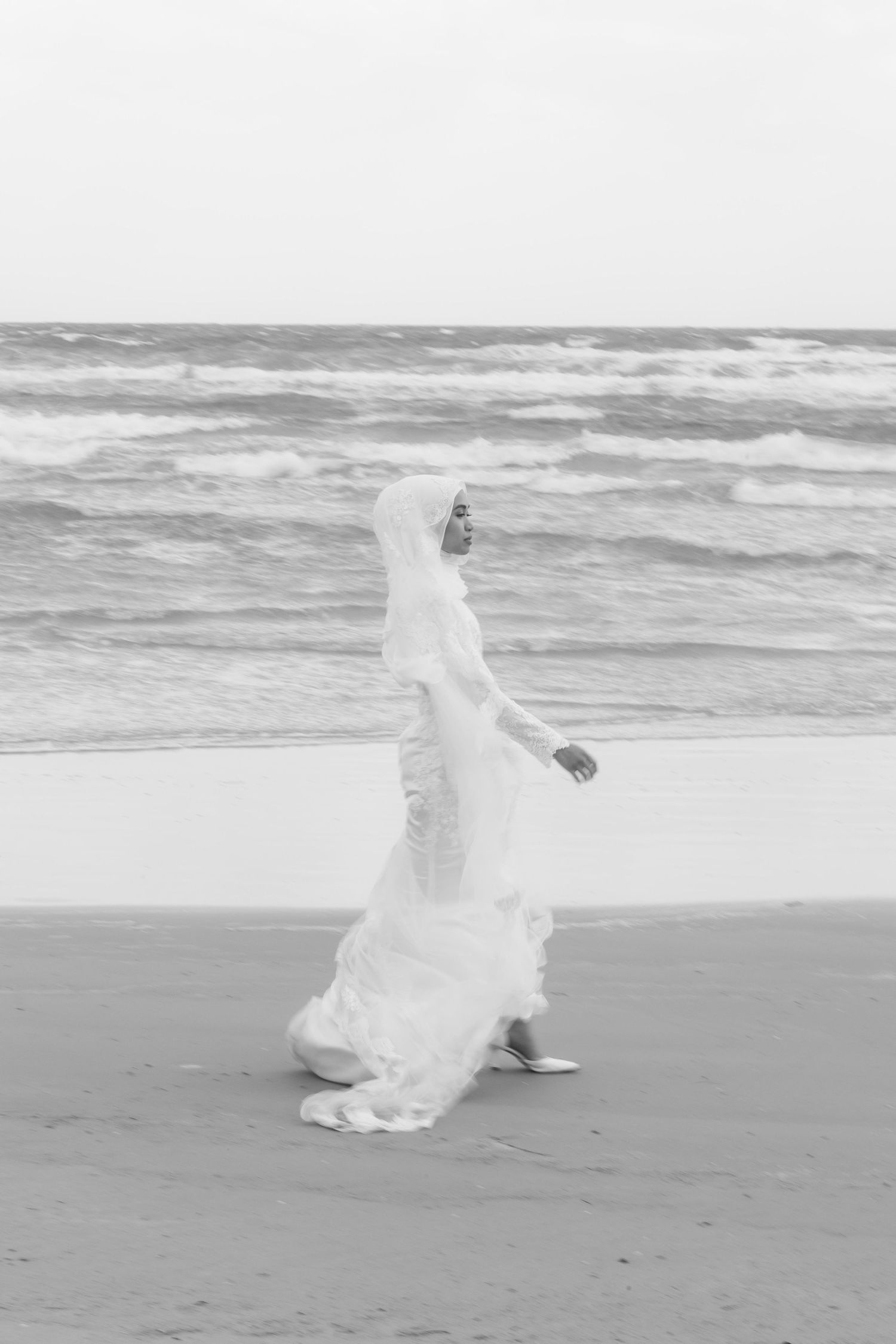 Black and white photo of Muslim bride in flowing white gown and hijab walking along a windy beach.