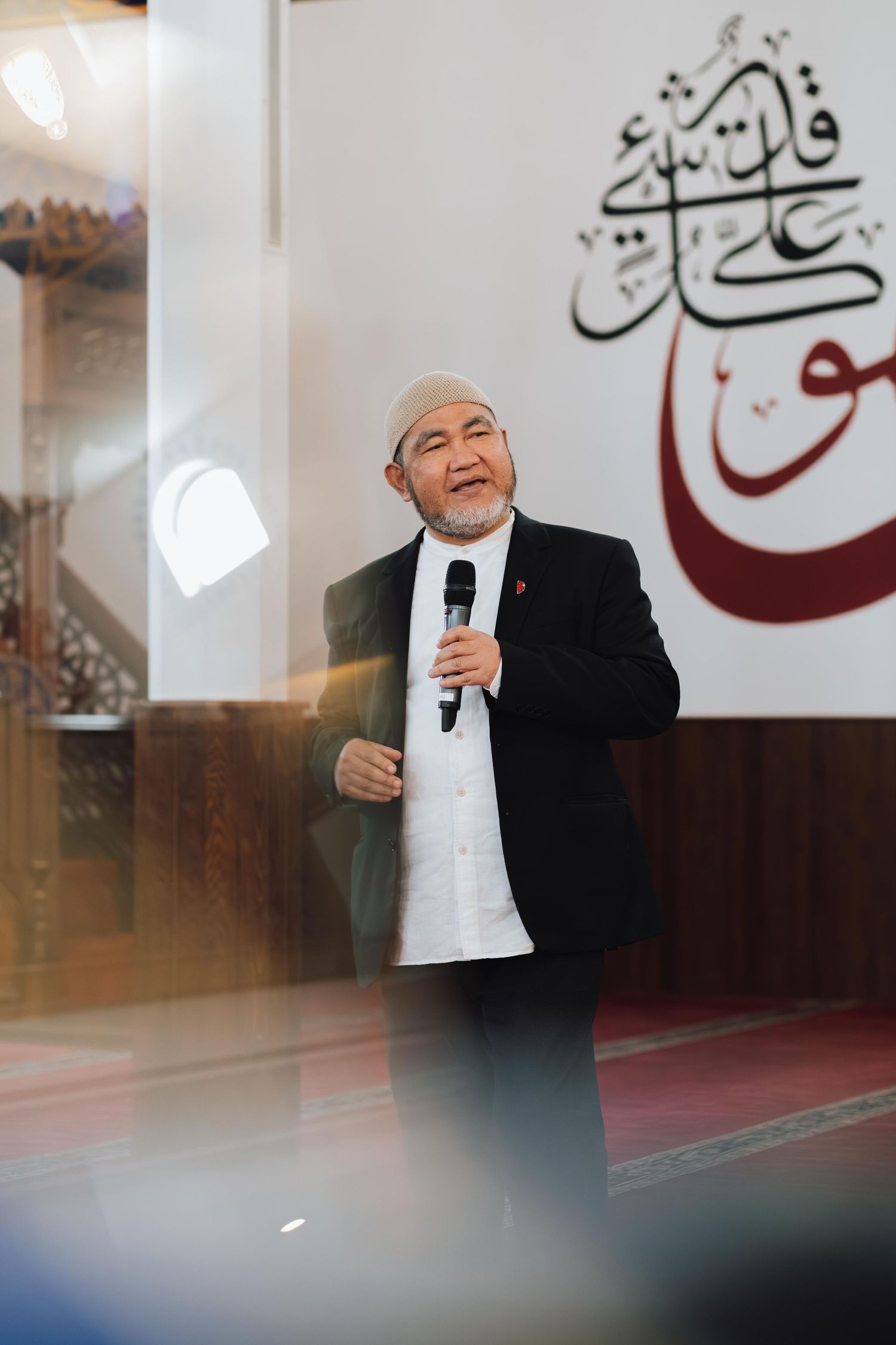 An elderly Muslim man in a black suit and white kufi cap speaks into a microphone inside a mosque.