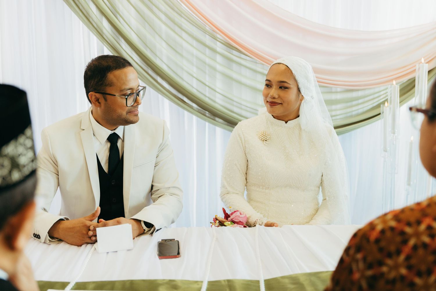 Muslim bride in white hijab and groom in white suit sit together at wedding ceremony.