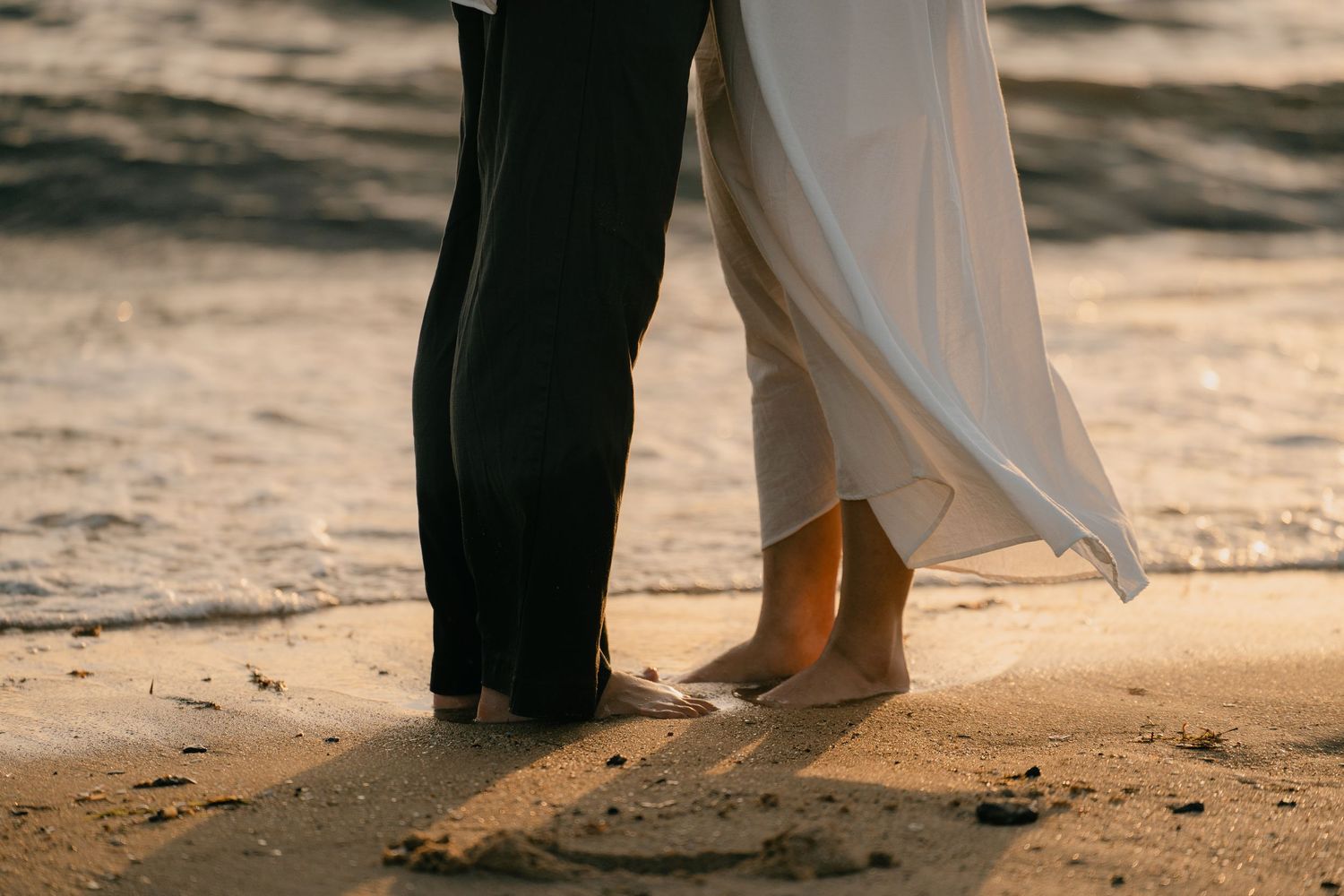 Barefoot couple standing together on a sandy beach at sunset, wearing wedding attire near ocean waves.