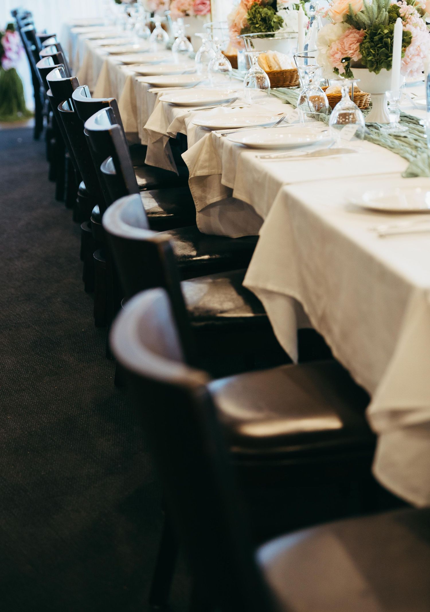Elegant wedding reception long table set with white linen, dark wooden chairs, floral centerpieces and candles.