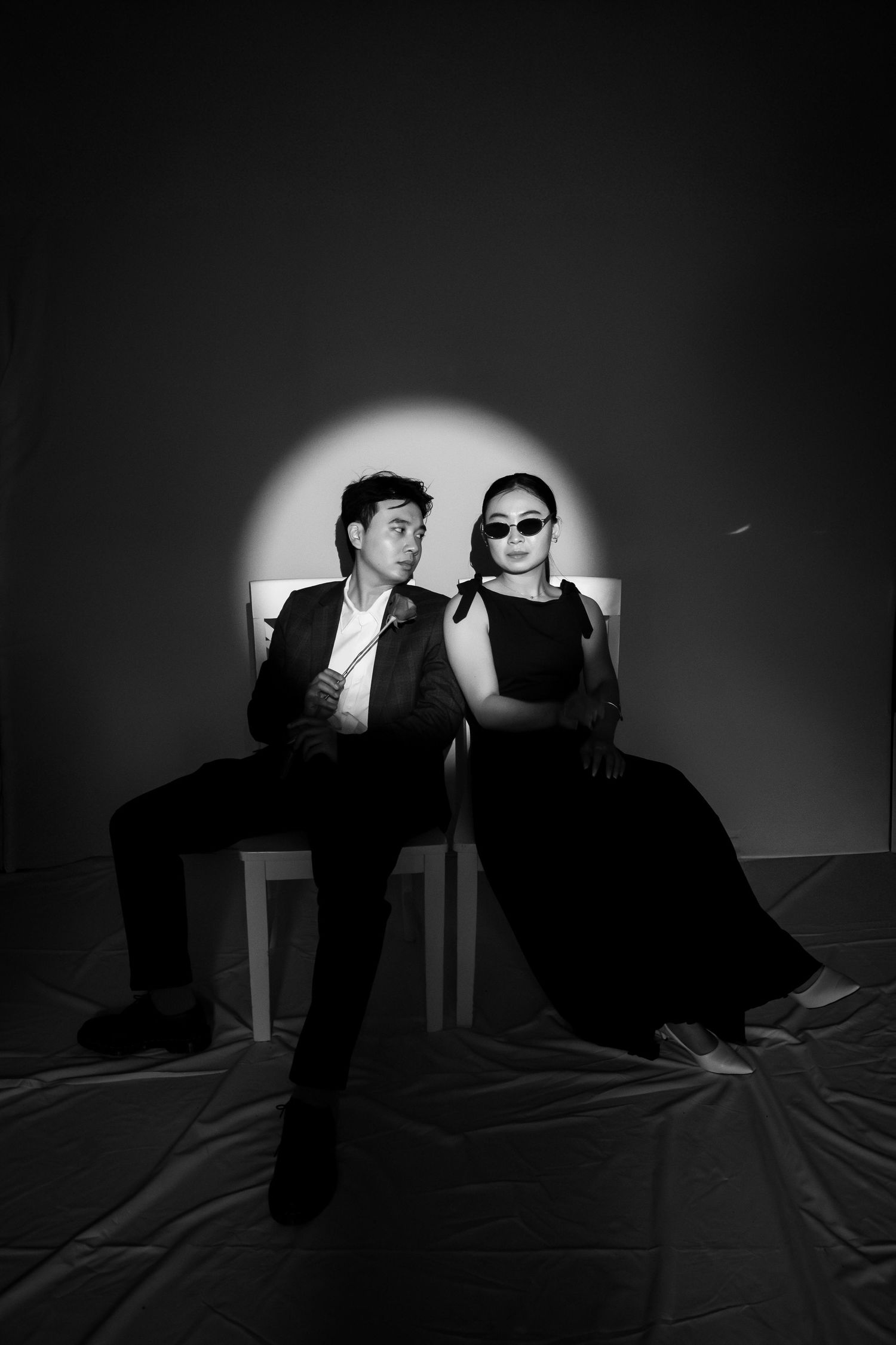 Black and white portrait of a man in a suit and woman in sunglasses seated together under a spotlight.