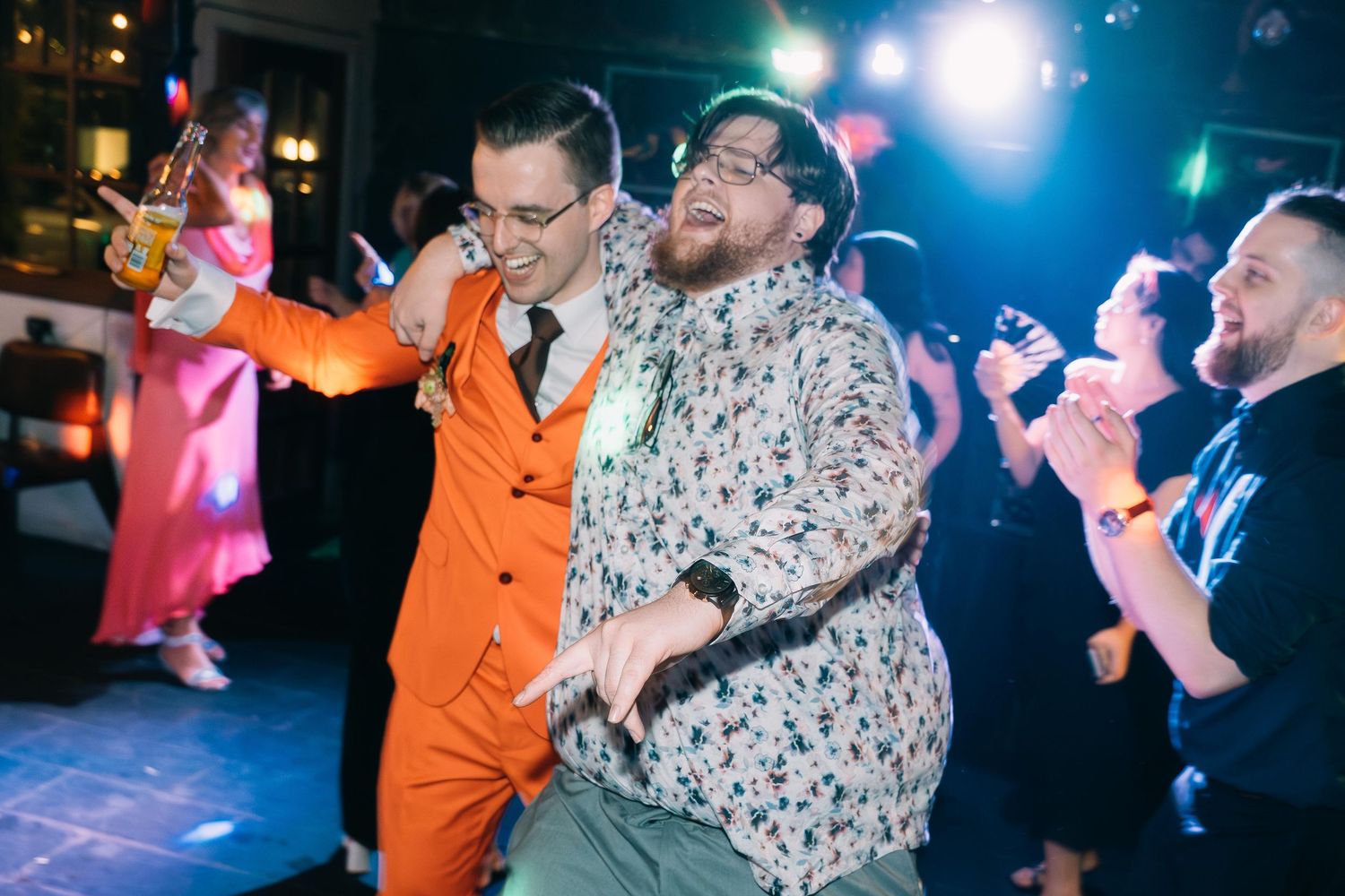 A groom in an orange suit dances energetically with a guest at a lively wedding reception.