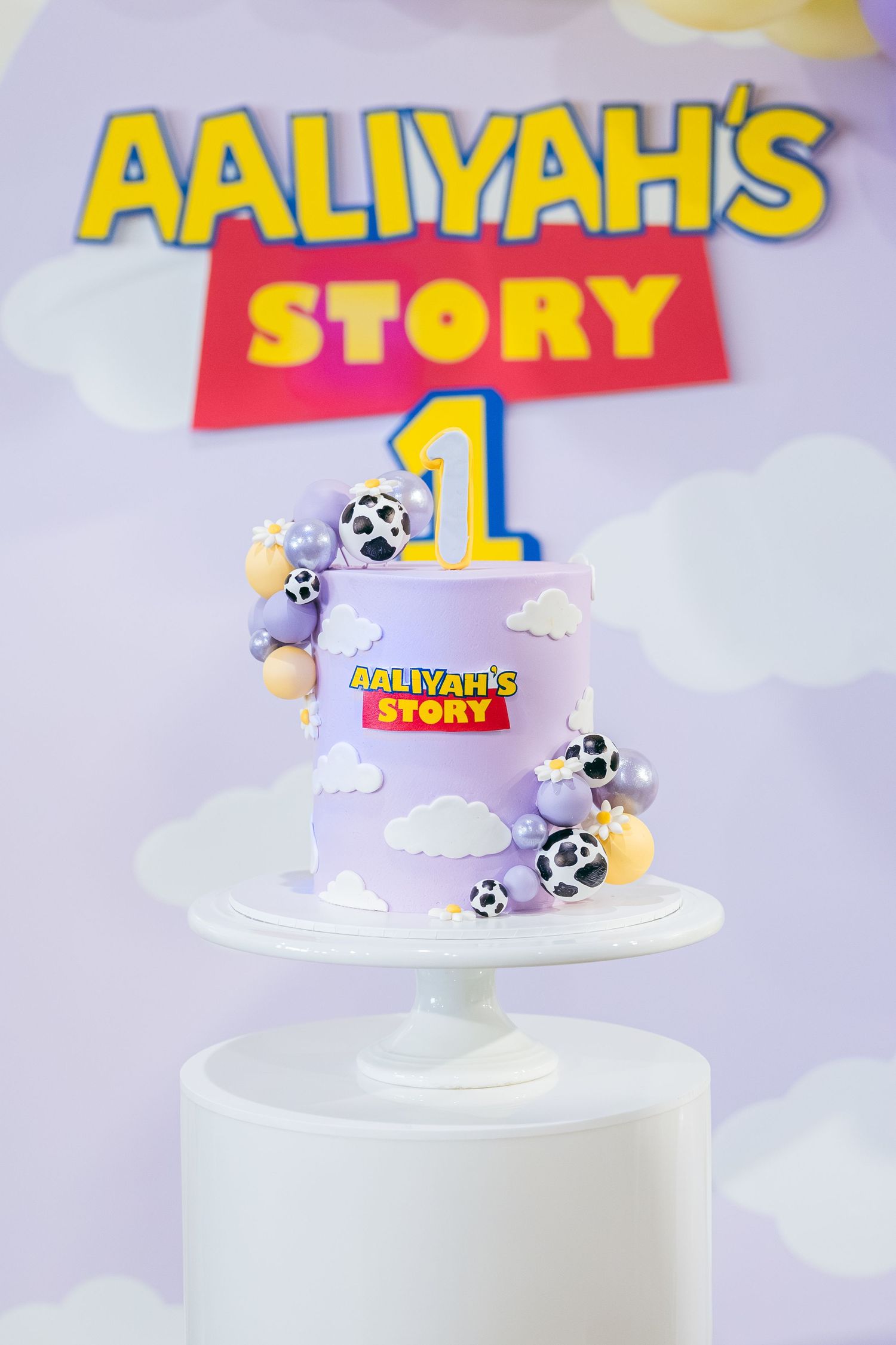 Purple and white birthday cake with Toy Story inspired design and cow print balloon decorations on a white pedestal stand.