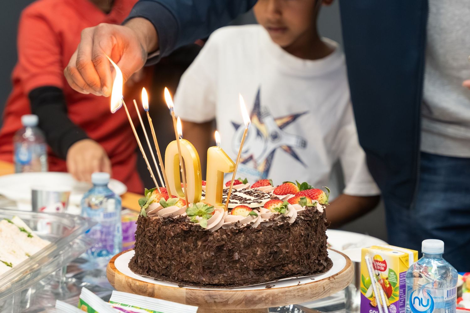 A chocolate birthday cake with lit candles surrounded by colorful decorations and party supplies on a festive table.