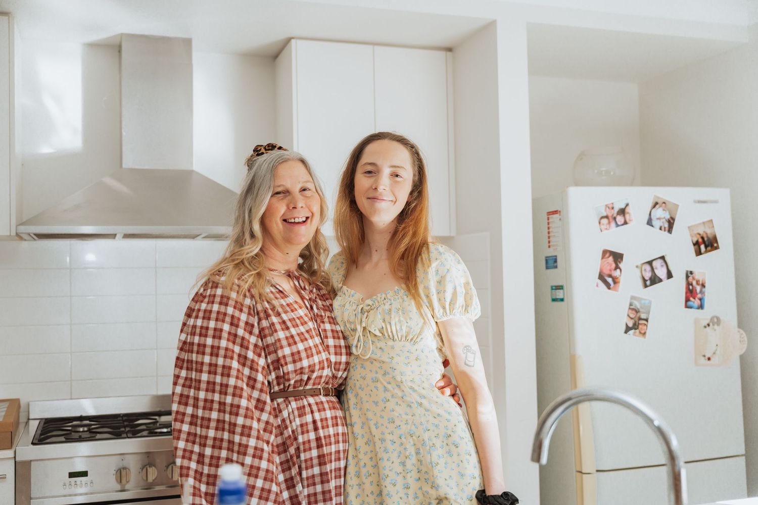 Two people laugh together in a bright white kitchen with a refrigerator and photos displayed on the wall.