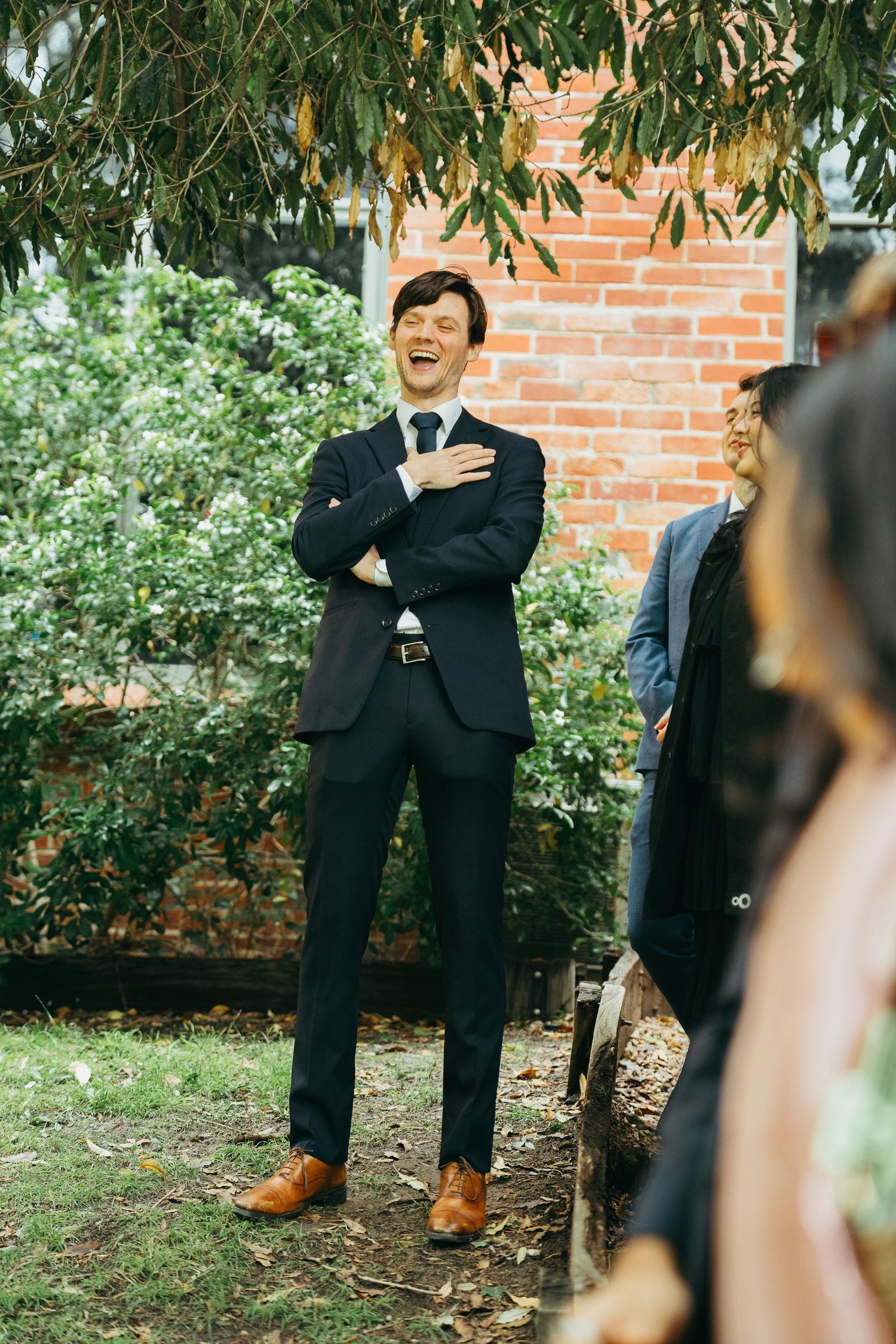 A groom in a navy suit laughs with hand on chest during an outdoor wedding ceremony.