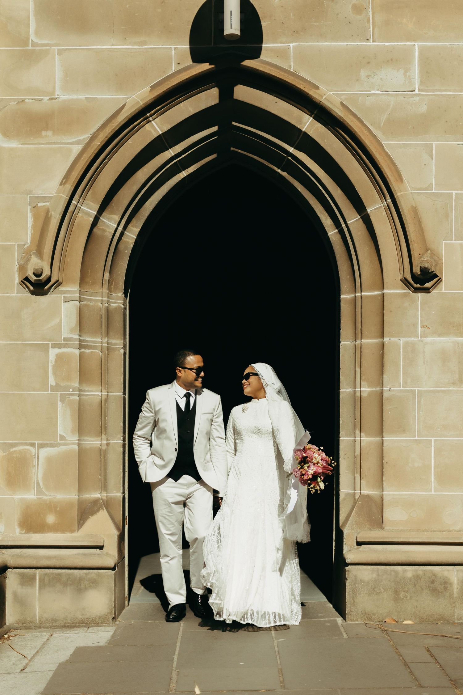 Muslim bride and groom in white suits posing under Gothic stone arch doorway on wedding day.