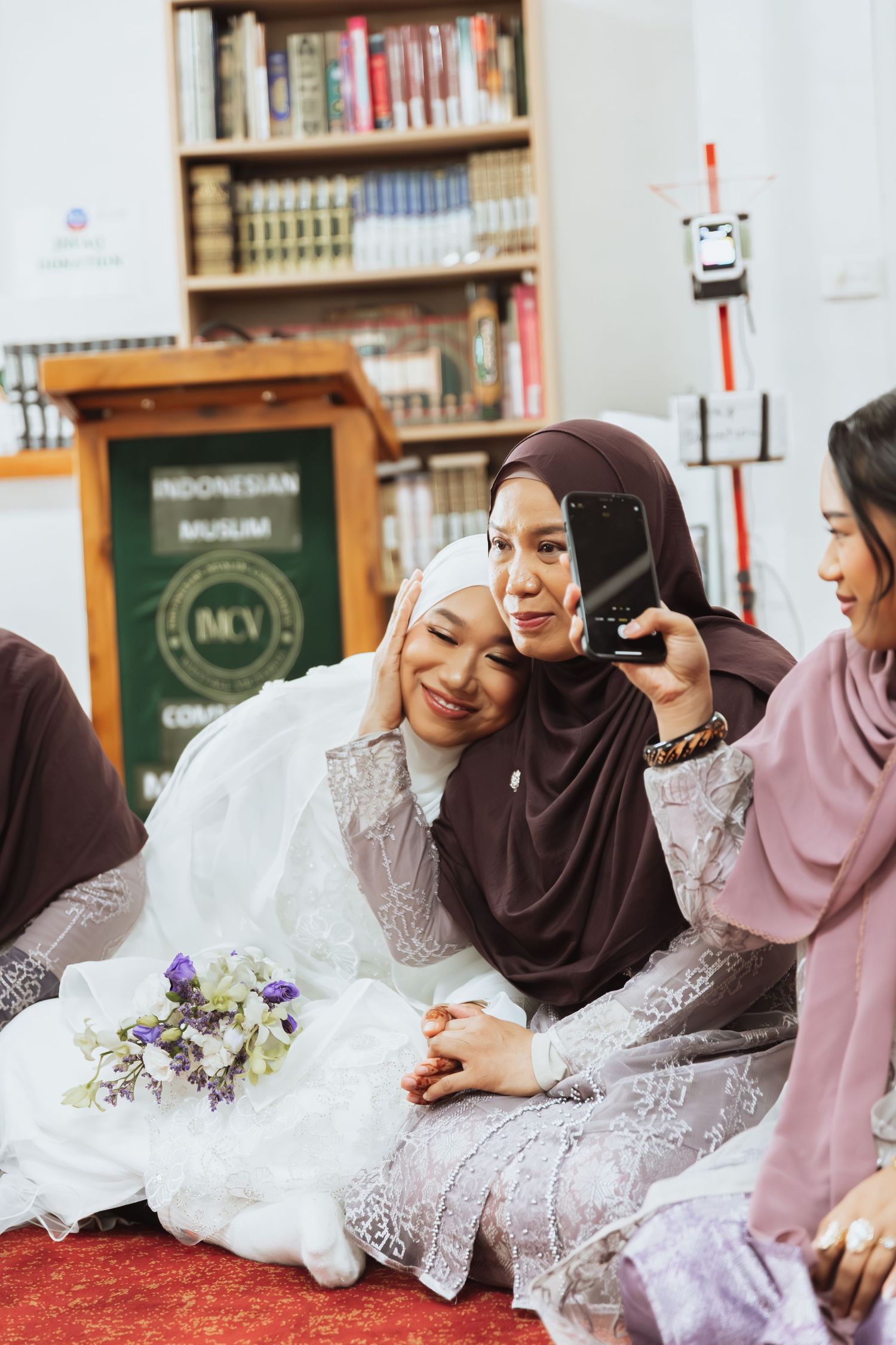 Muslim women in hijabs taking a selfie with a bride in white at an Indonesian Muslim community wedding ceremony.