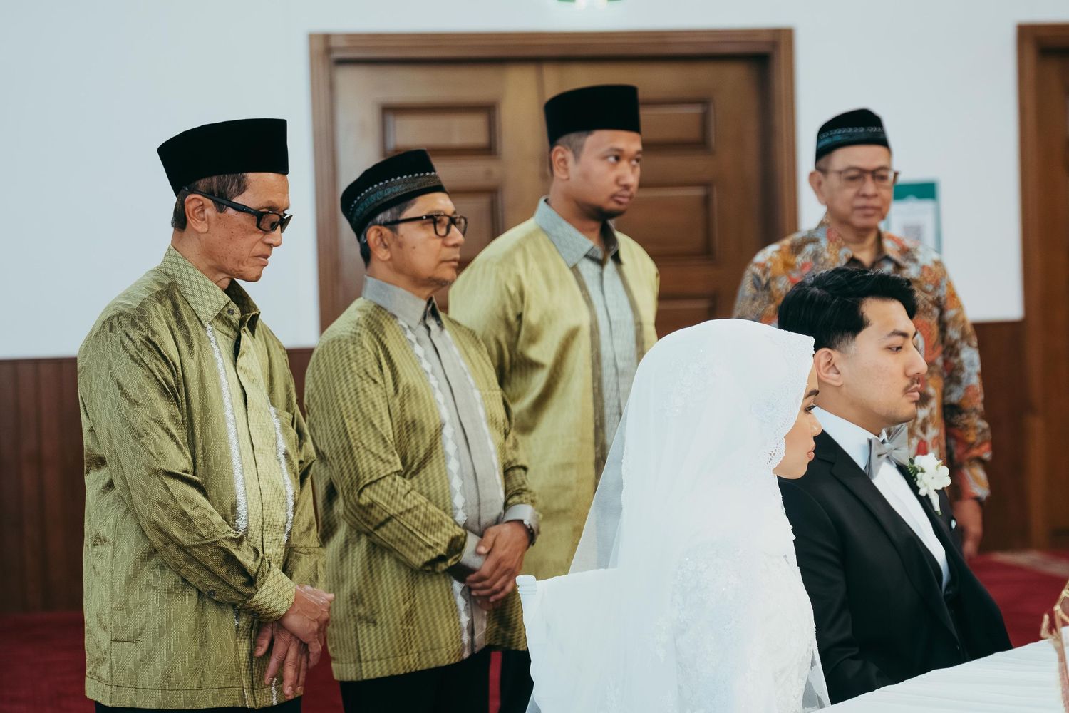 Muslim men in traditional golden baju kurung standing solemnly during an Islamic wedding ceremony indoors.