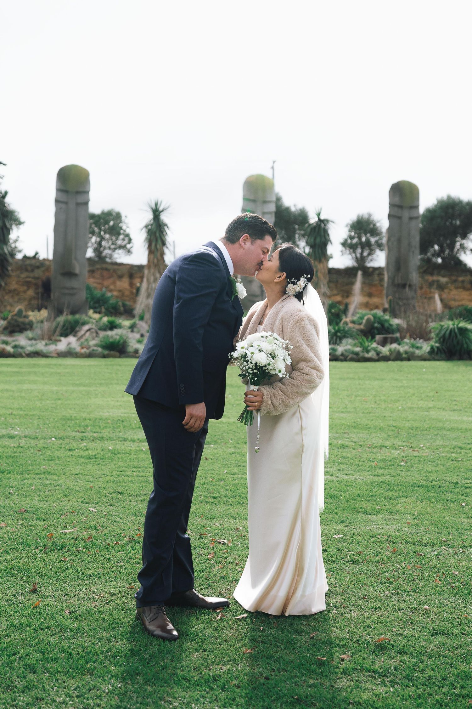 Bride and groom share a kiss on green grass with stone garden sculptures and coastal cliffs in background.