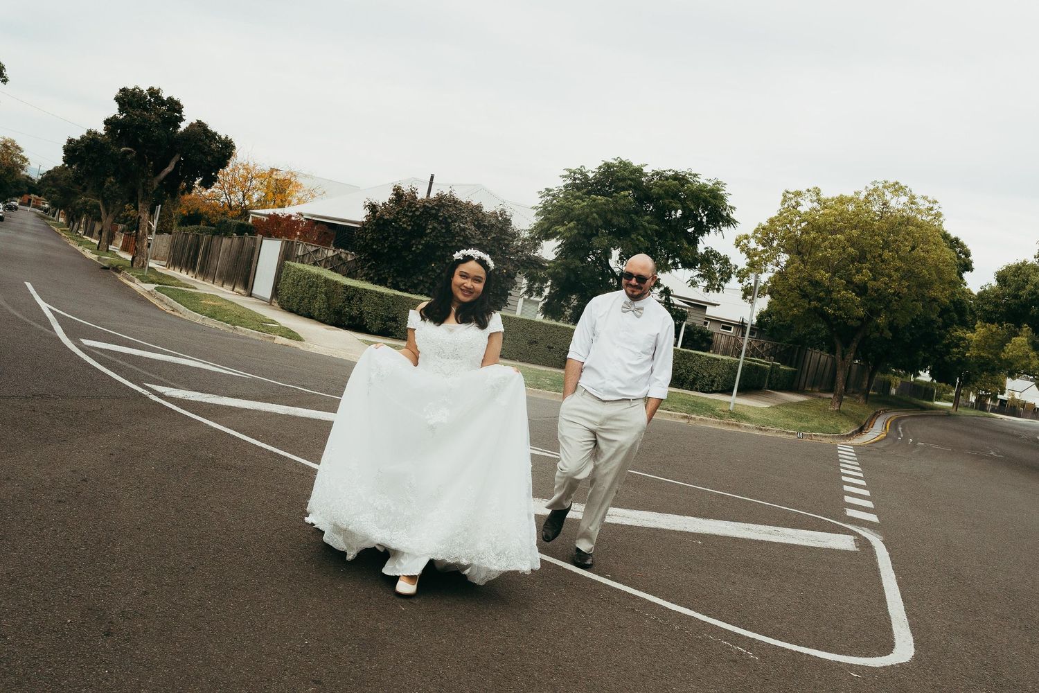Bride and groom walking joyfully across a suburban street, bride in white gown, groom in grey suit with sunglasses.