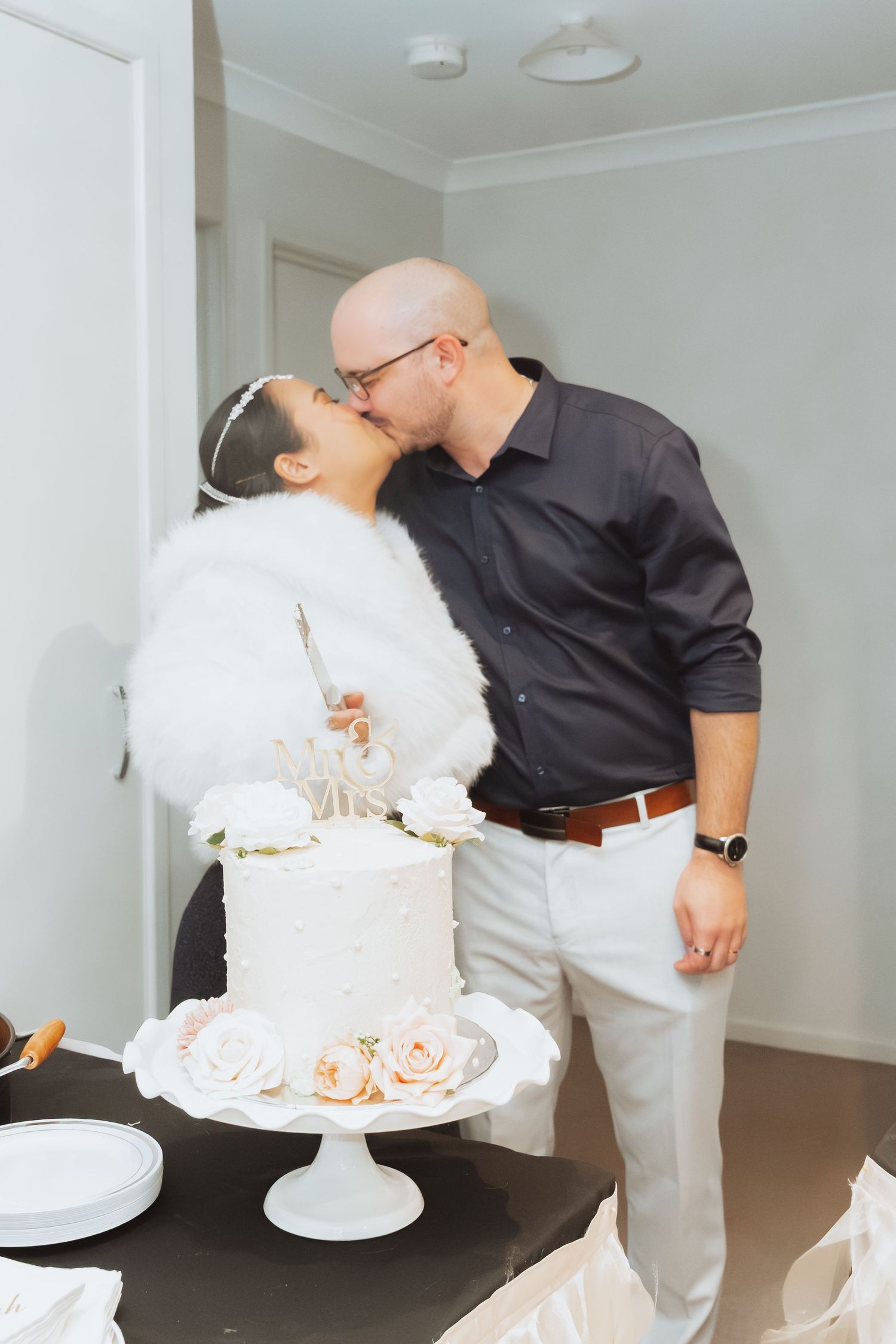 A couple shares a romantic moment while cutting their elegant white wedding cake at an indoor celebration.