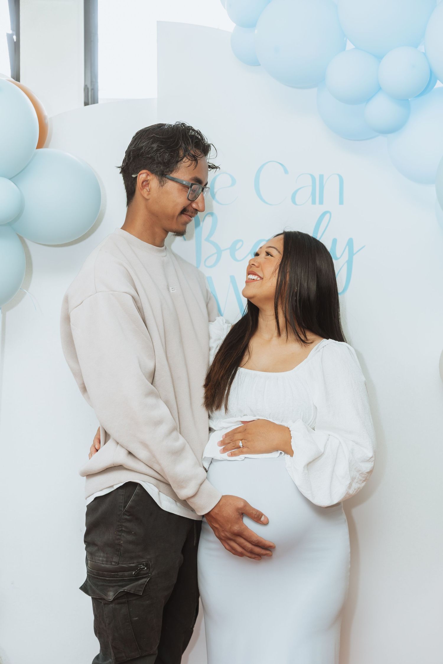 Couple in white and gray outfits embracing during pregnancy photoshoot with blue cloud backdrop.