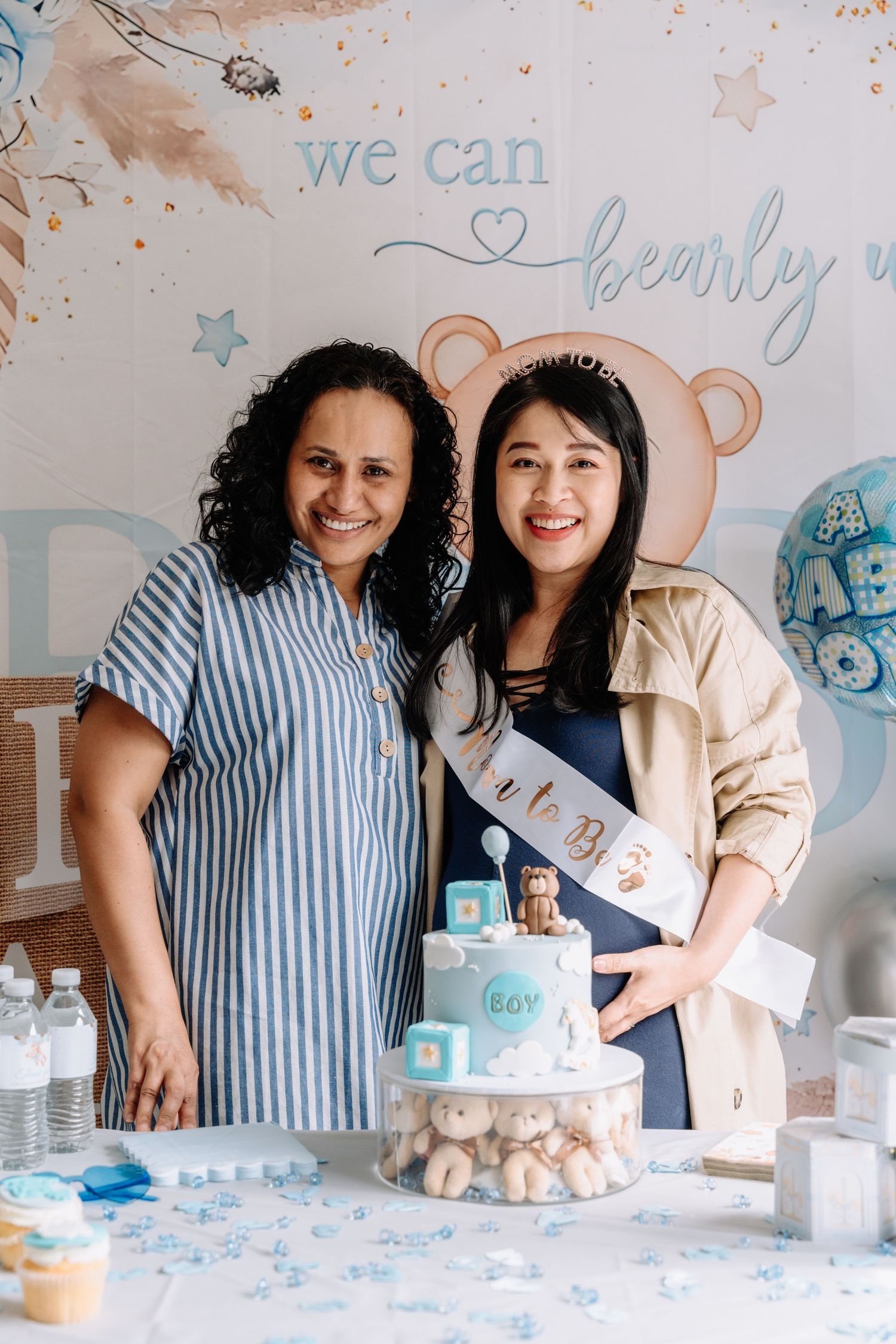 Two guests pose together by a decorated dessert table with a blue and white teddy bear themed cake.