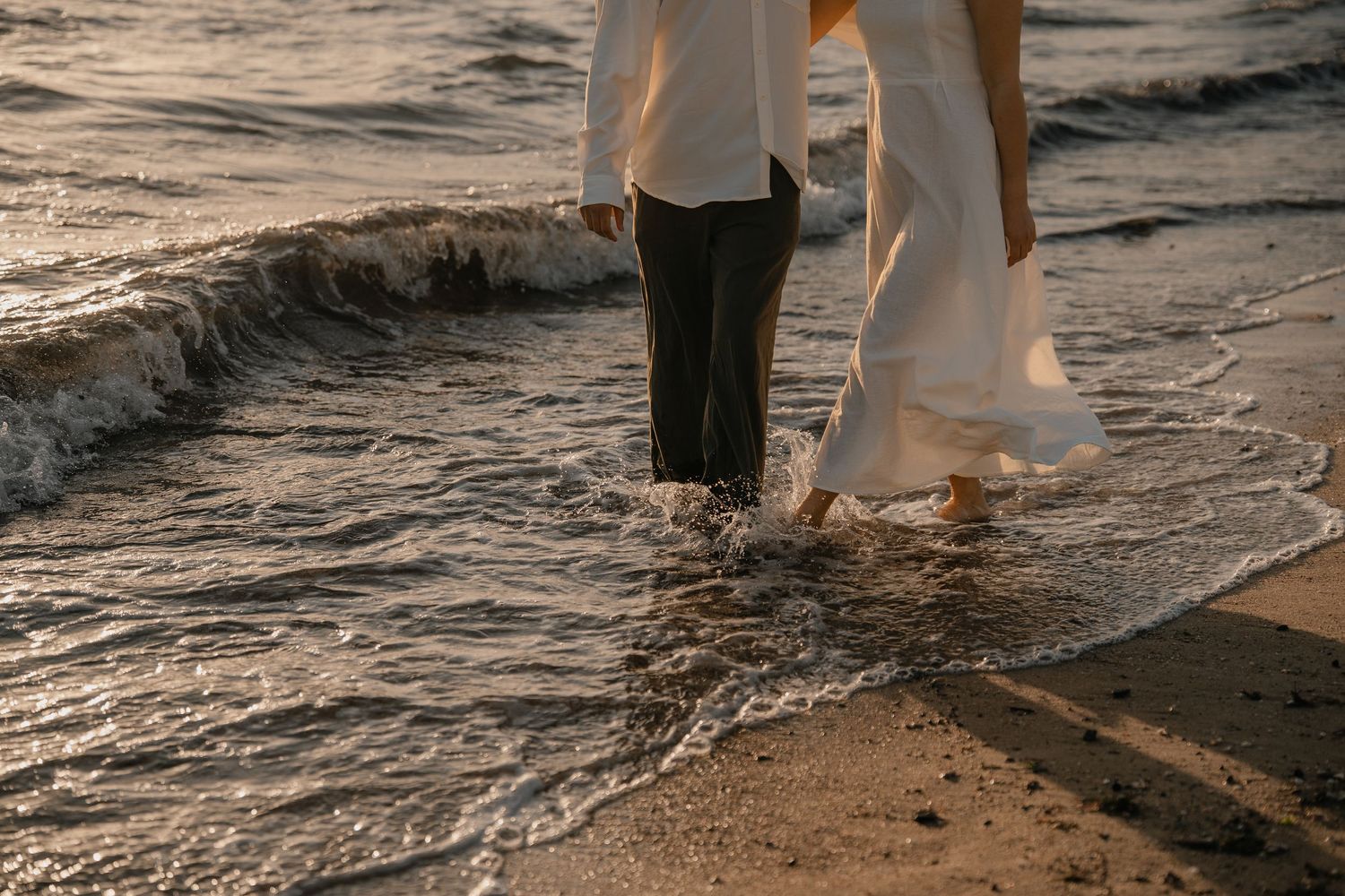 Couple walking barefoot on sandy beach shore at sunset in wedding attire, waves washing over their feet.