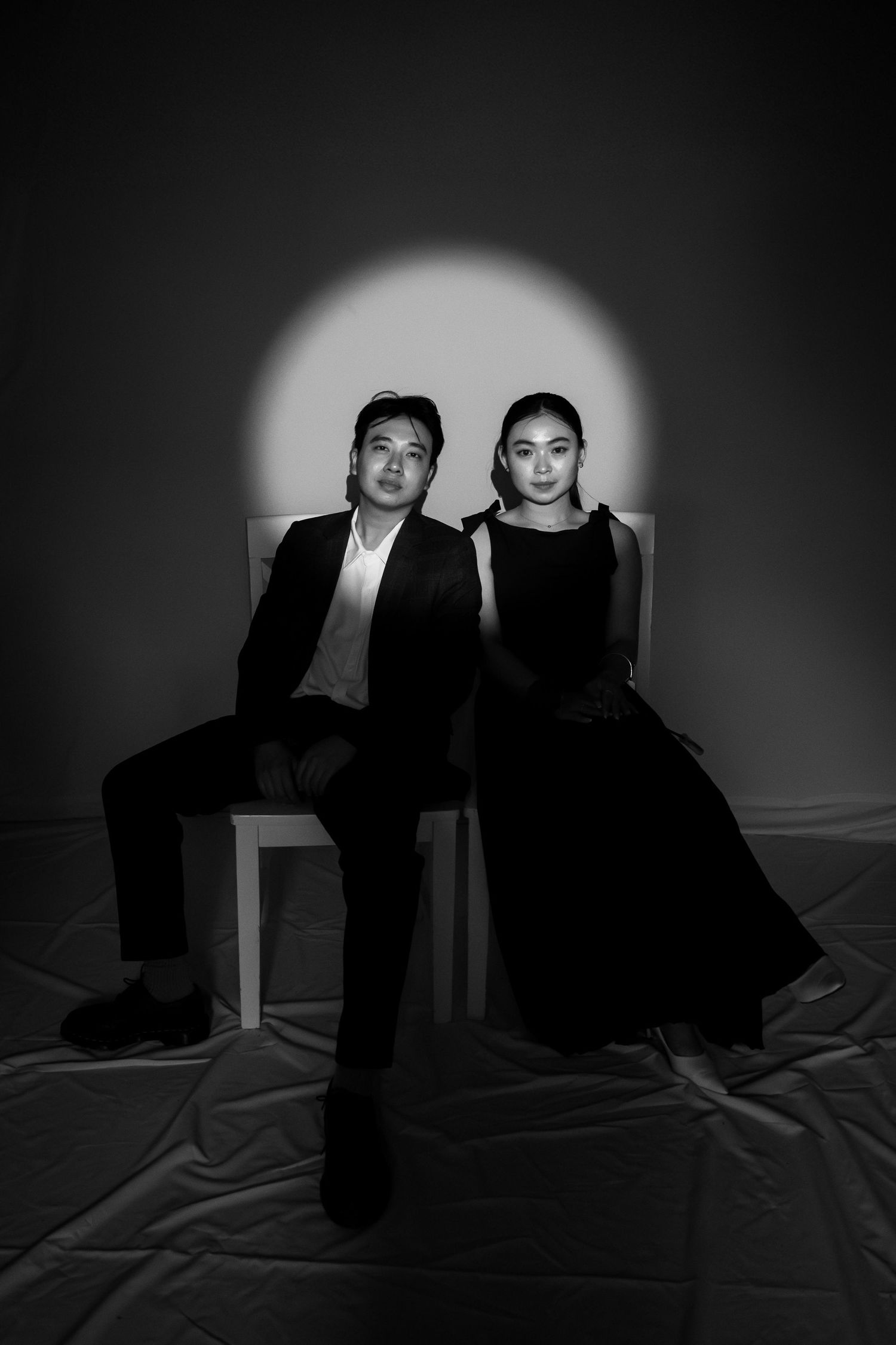 Black and white photo of a man and woman in formal attire seated against a dark backdrop with circular spotlight.