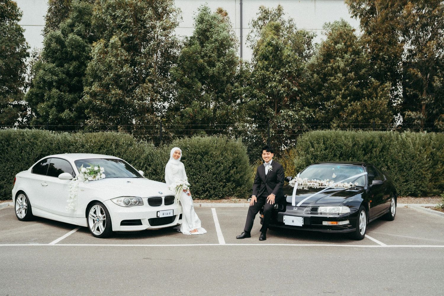 Muslim bride and groom pose between a white BMW and black car in a parking lot.