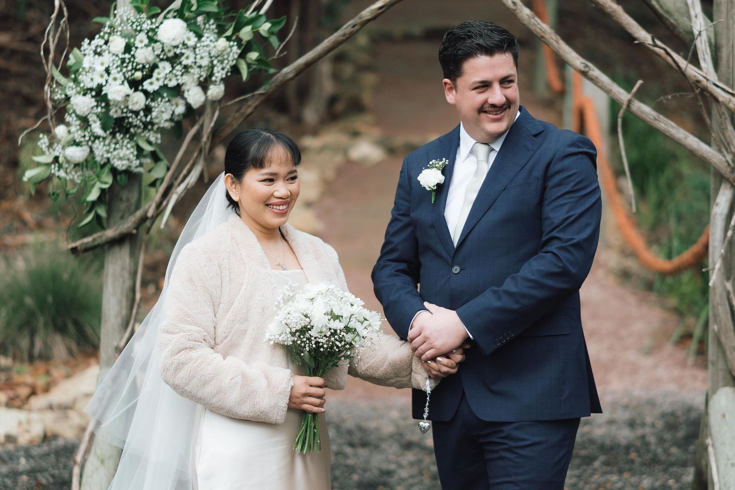 Smiling bride and groom holding hands at outdoor wedding ceremony under rustic floral arch.