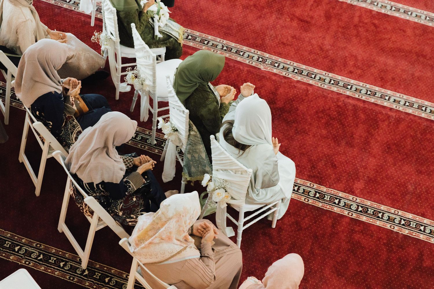 Guests in hijabs seated on white chairs over red carpet at Muslim wedding ceremony.