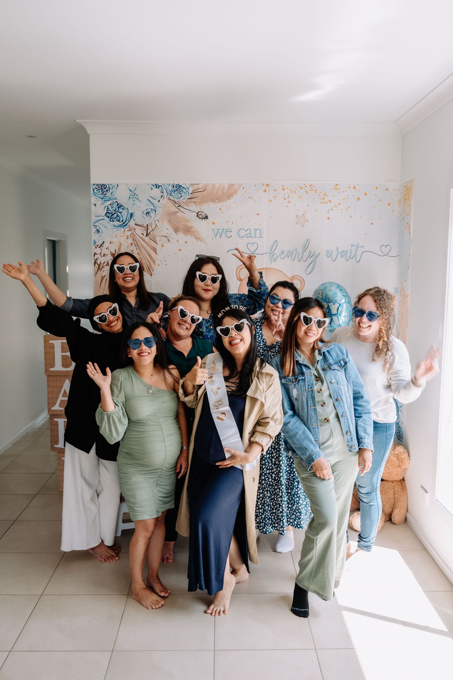 A group of friends pose together wearing sunglasses against a floral wall backdrop at a party.