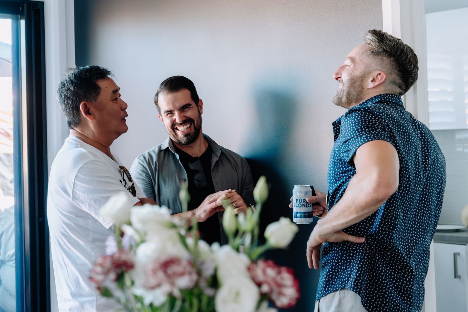 Three friends laughing together at a social gathering with flowers in the foreground.