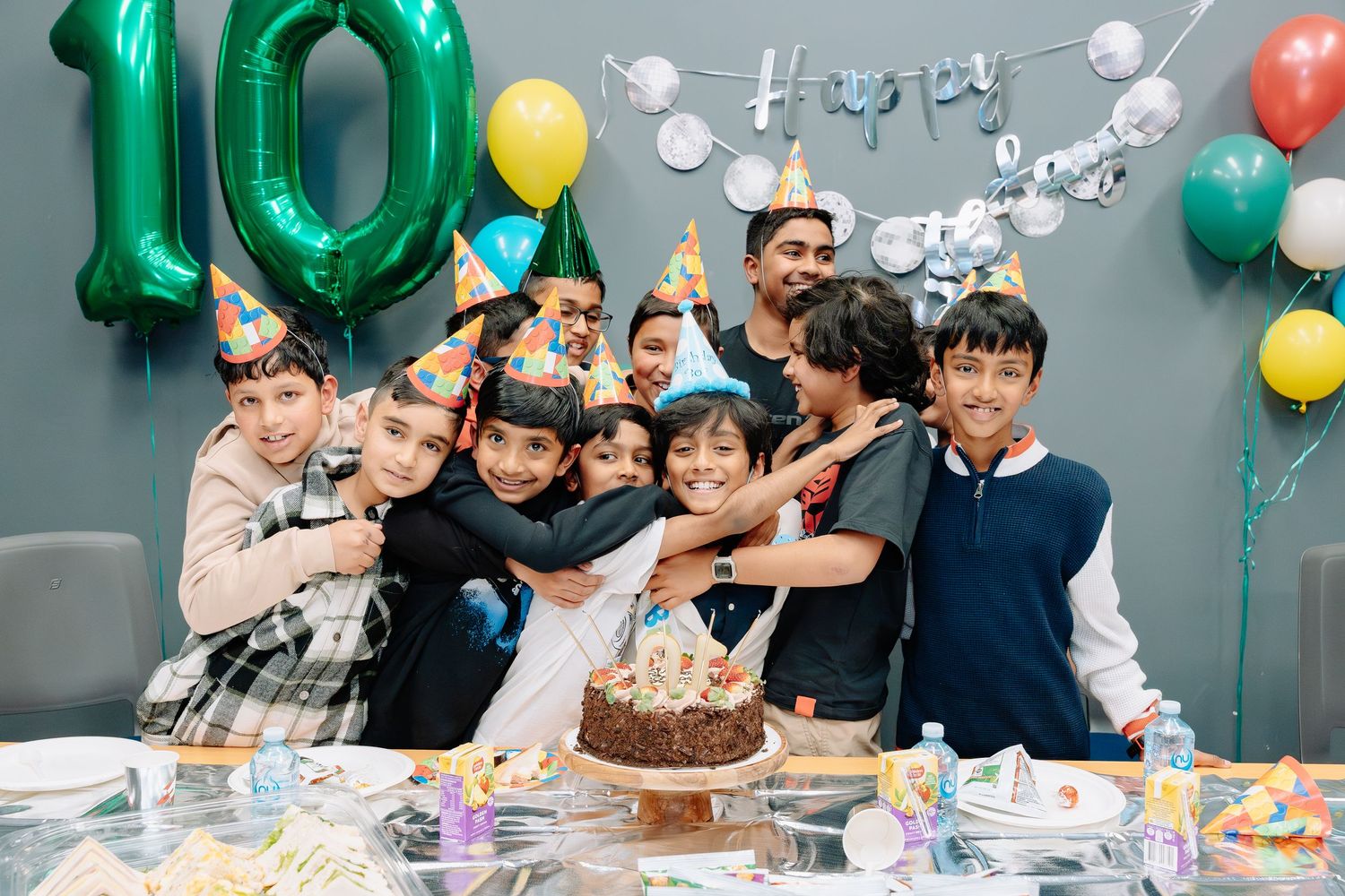 A group celebrates at a party with colorful balloons and decorations while gathered around a birthday cake.