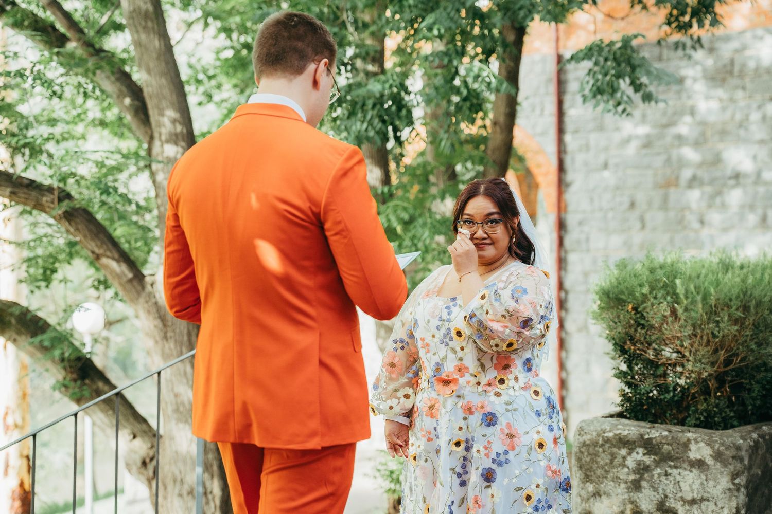 Groom in vibrant orange suit reads vows to smiling bride in floral dress at outdoor garden wedding.