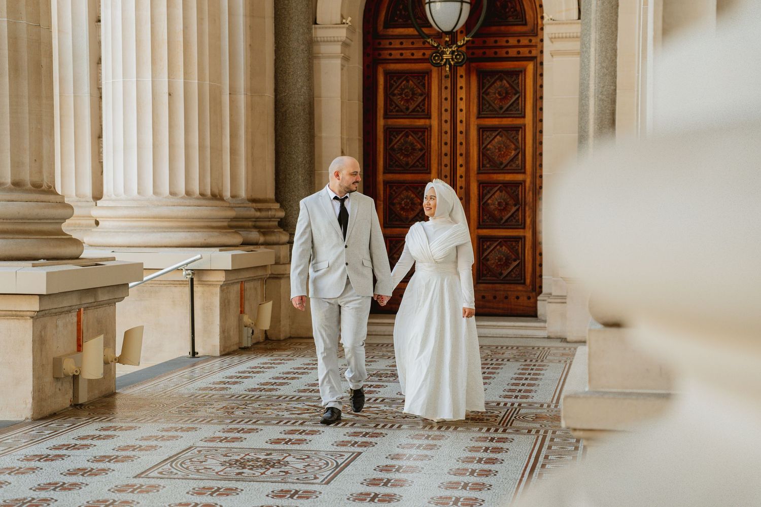 Muslim bride and groom in white wedding attire walk hand in hand through elegant columned hall with ornate wooden doors.