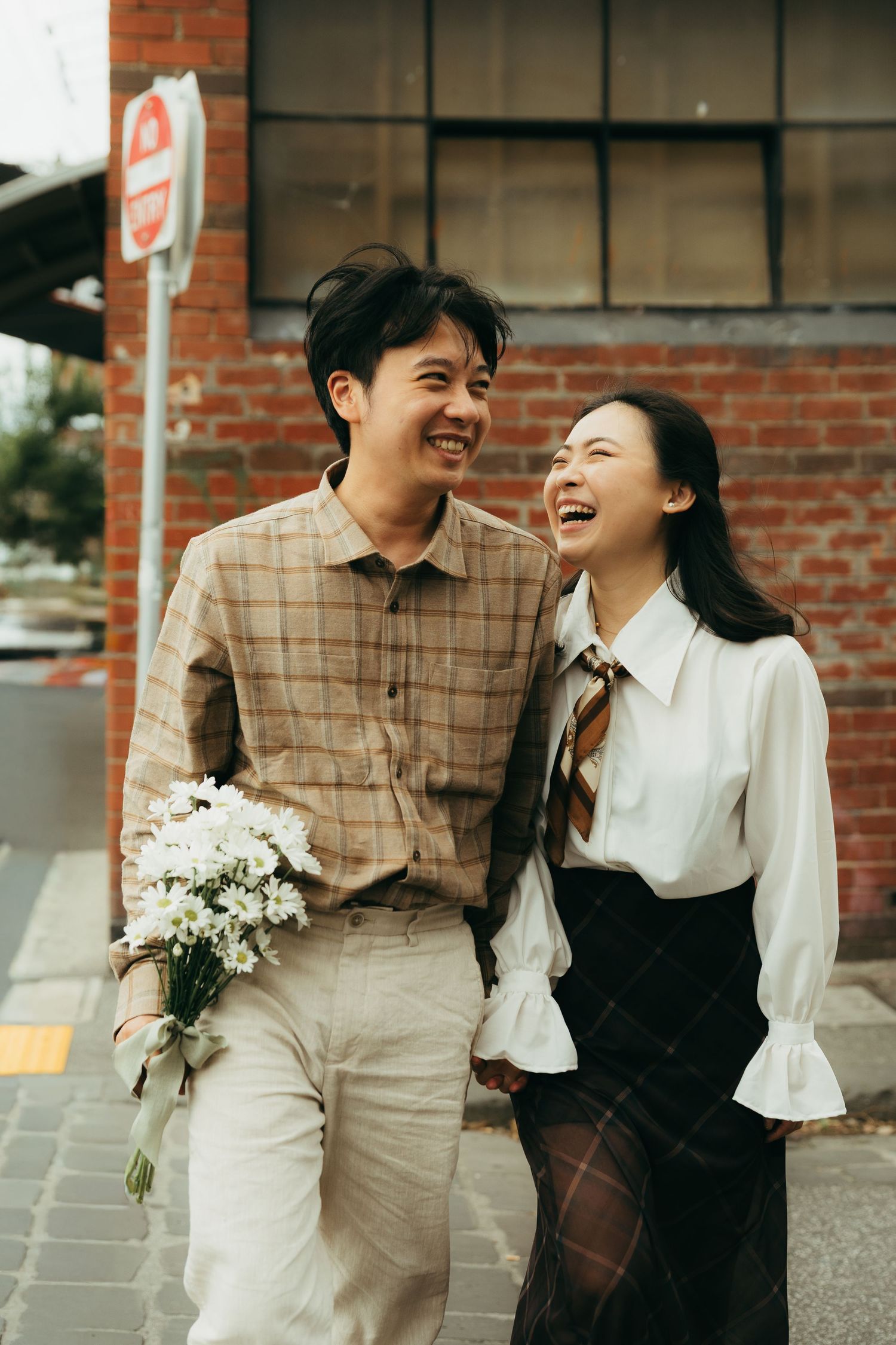 Happy Asian couple walking together, man holding white flowers, laughing outdoors near brick wall.