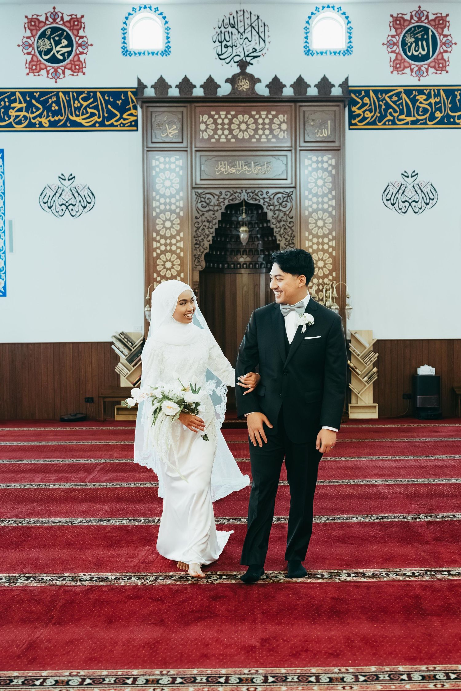 Muslim bride in white hijab and groom in black suit smile while standing in decorated mosque at wedding.